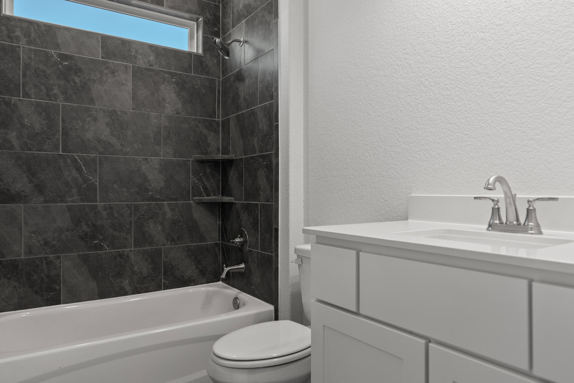 Bathroom with white bathtub against tiled wall, white toilet with closed lid, sink with metal faucet, shower area featuring window with blue sky visible, mirror above sink, neutral