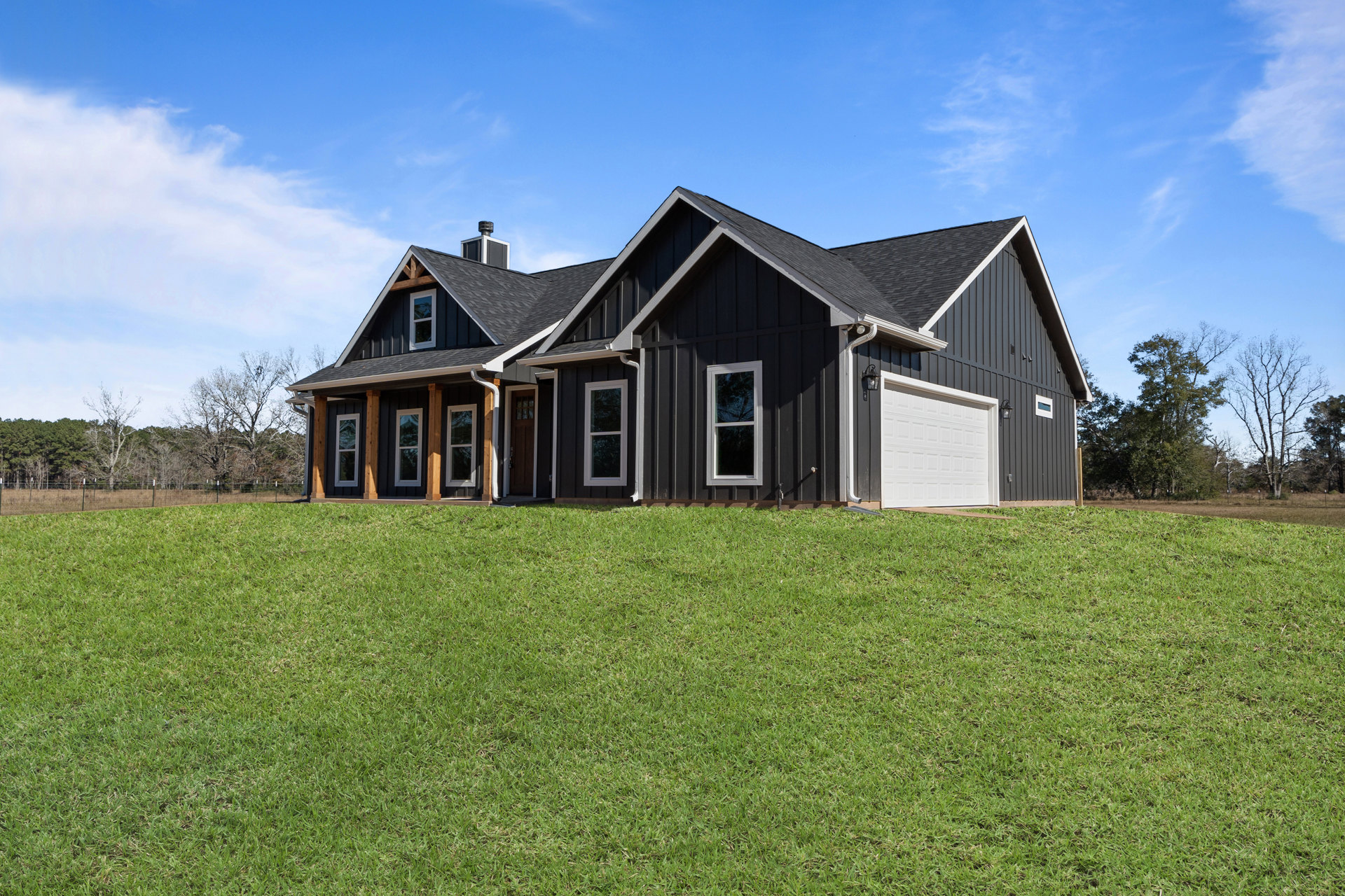 Modern black siding house with white framed windows, expansive green lawn, mature trees, and clear blue sky