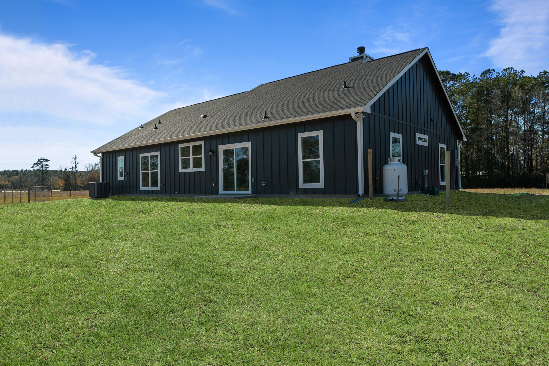 Two-story farmhouse with white siding, large windows with white frames, manicured green lawn, blue outdoor chair near the back, clear blue sky overhead, and a white water tank with