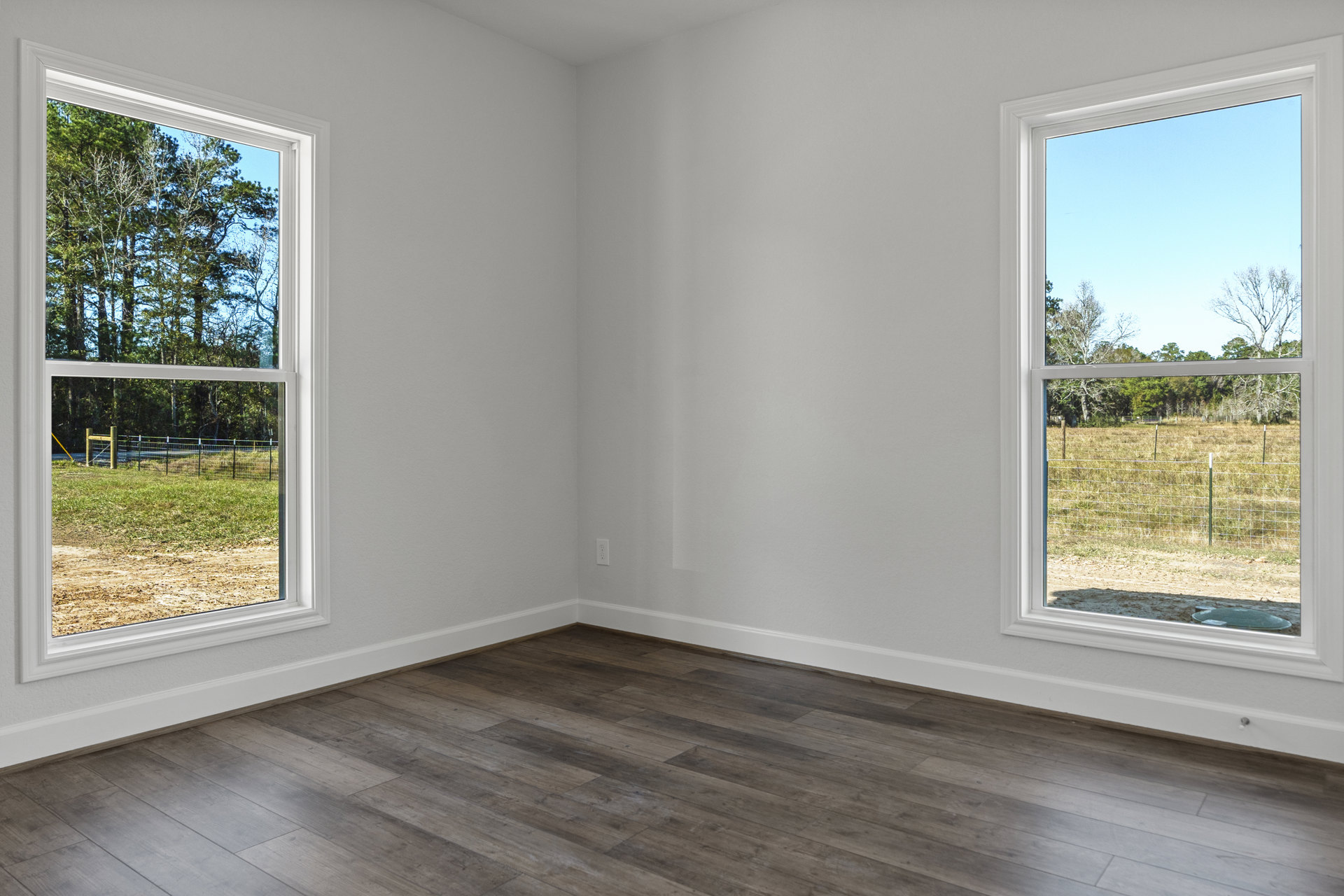Wood floor room with white walls, two large windows, view of trees and fence outside