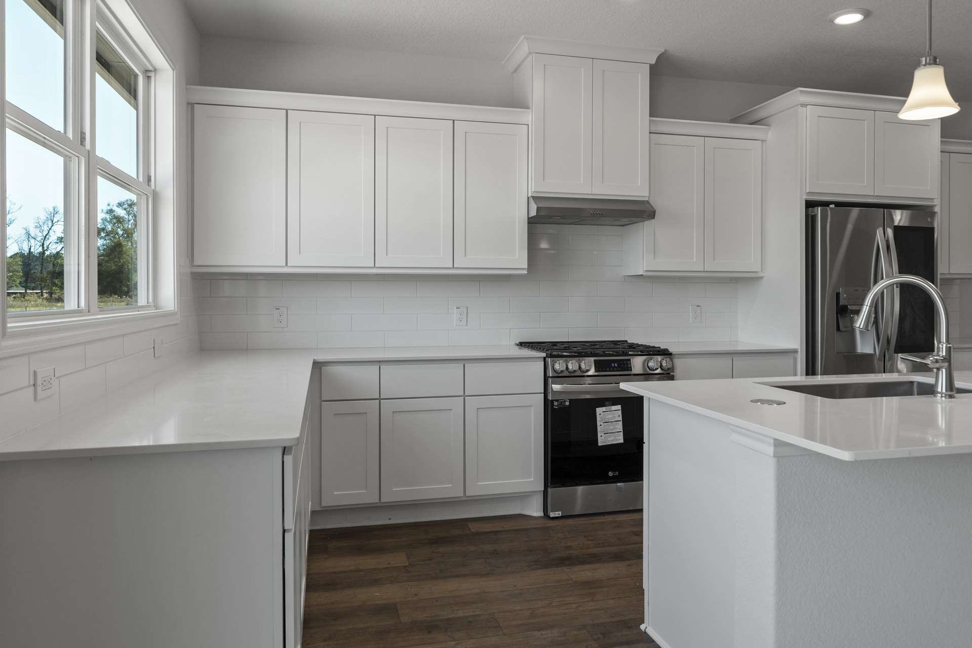 White kitchen with shaker cabinets, stainless steel stove and refrigerator, white island with undermount sink, wood flooring, white walls, and modern pendant light fixture.