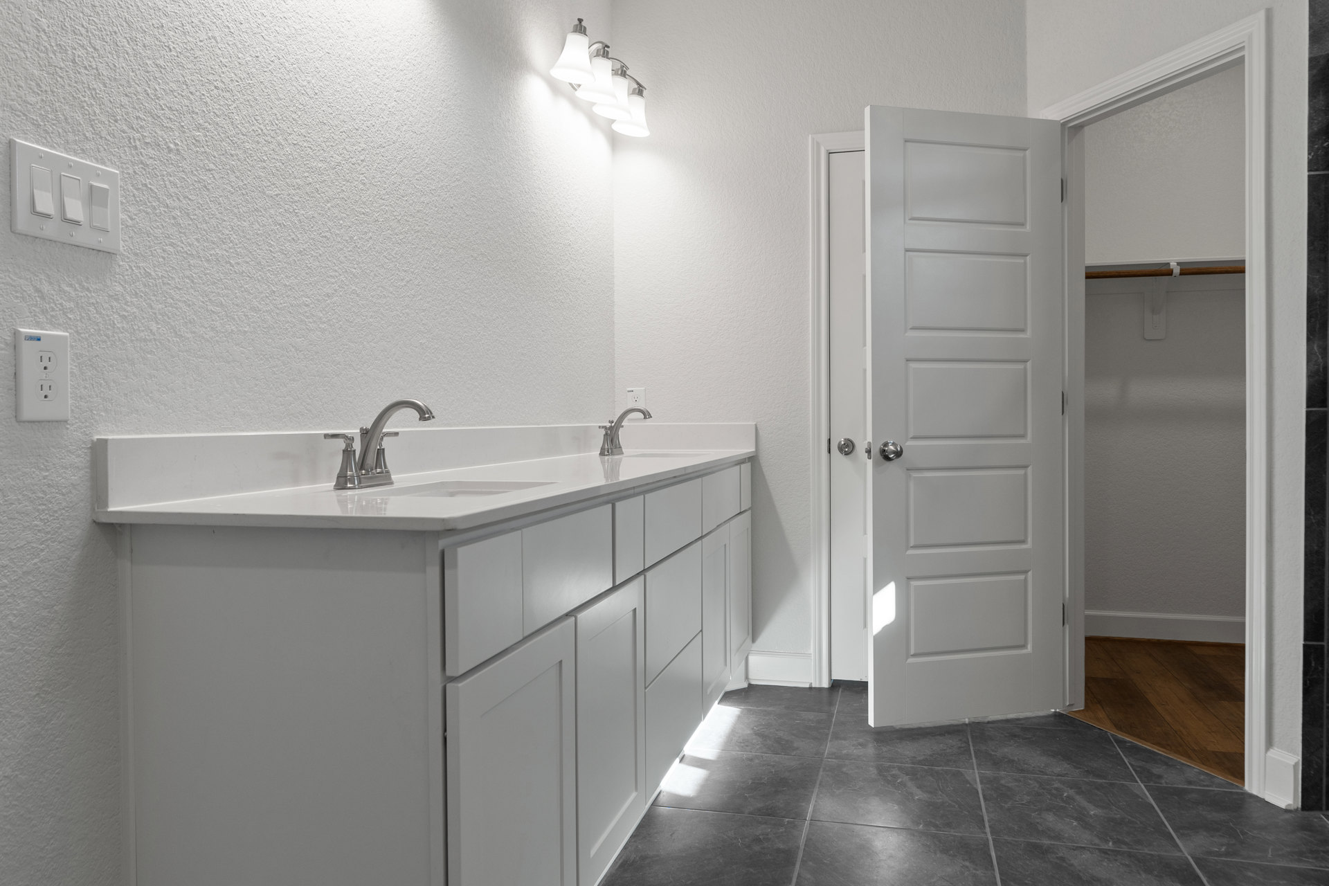 Bathroom with white cabinets, white walls, grey tile floor, white door with silver knobs, four-light fixture, close-up of light switch and electrical outlet