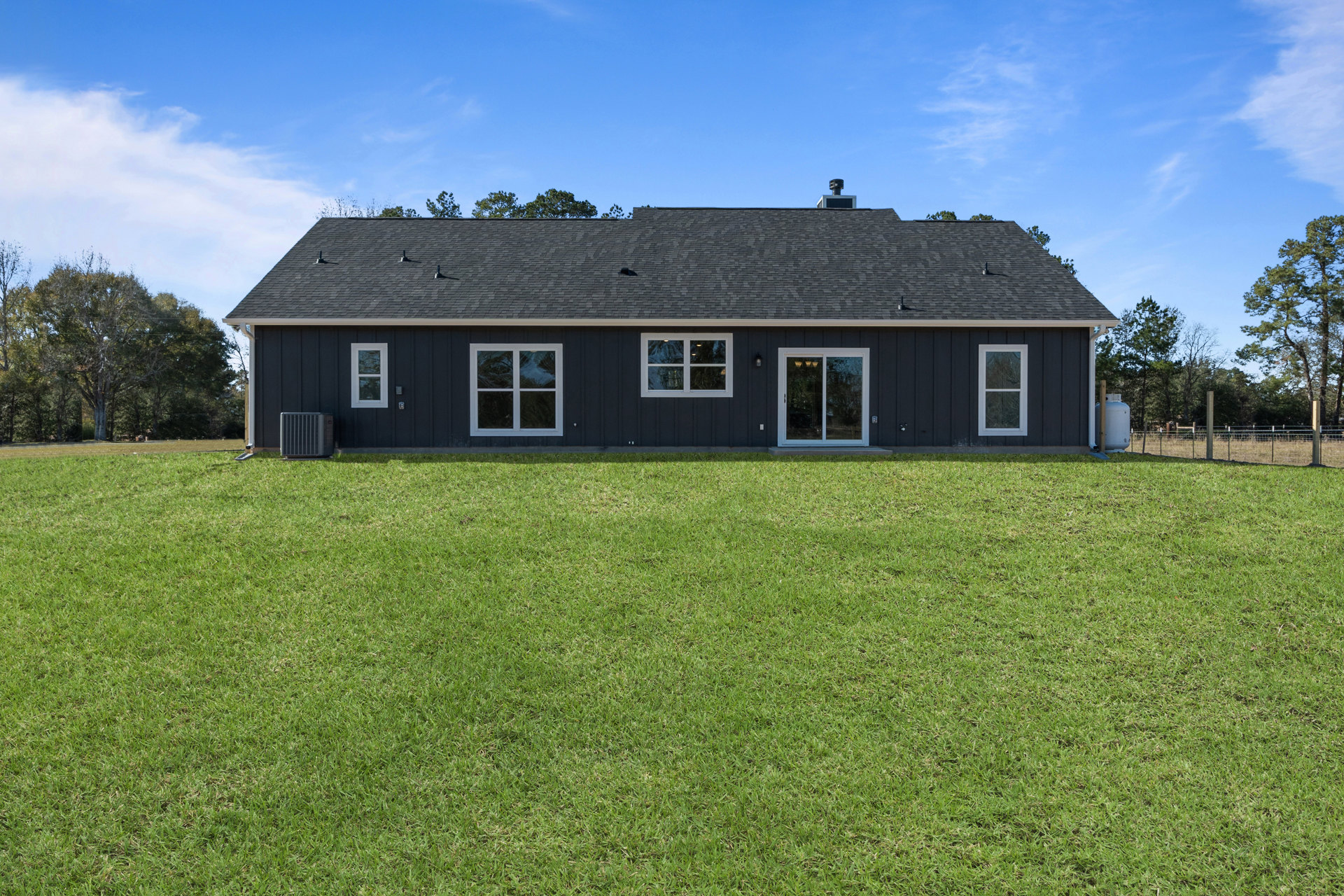 Two-story house with black roof, white-framed windows, white sliding door, chimney, and large metal HVAC unit, surrounded by green lawn and trees under cloudy sky