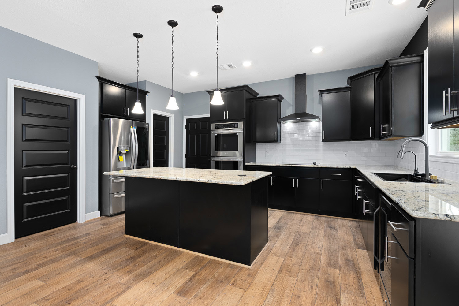 Black kitchen cabinets, marble island with waterfall edge, stainless steel oven, black door, marble countertop, built-in refrigerator, ceiling light fixture