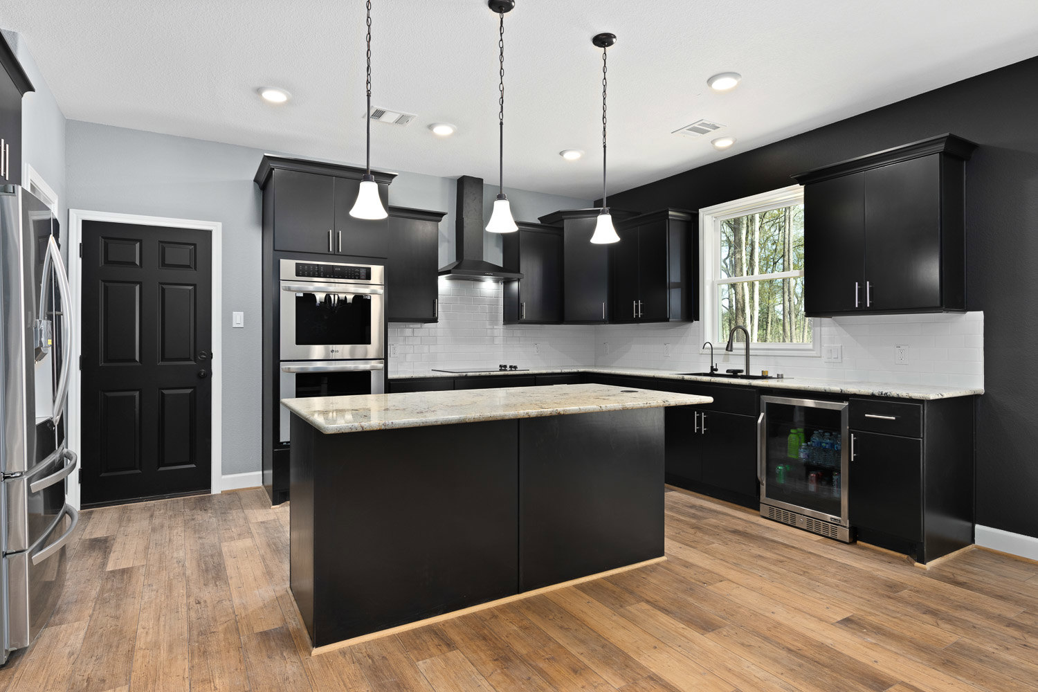 Black kitchen cabinets with marble countertops, wood flooring, stainless steel oven, and refrigerator stocked with soda bottles and cans; black door with white trim visible in
