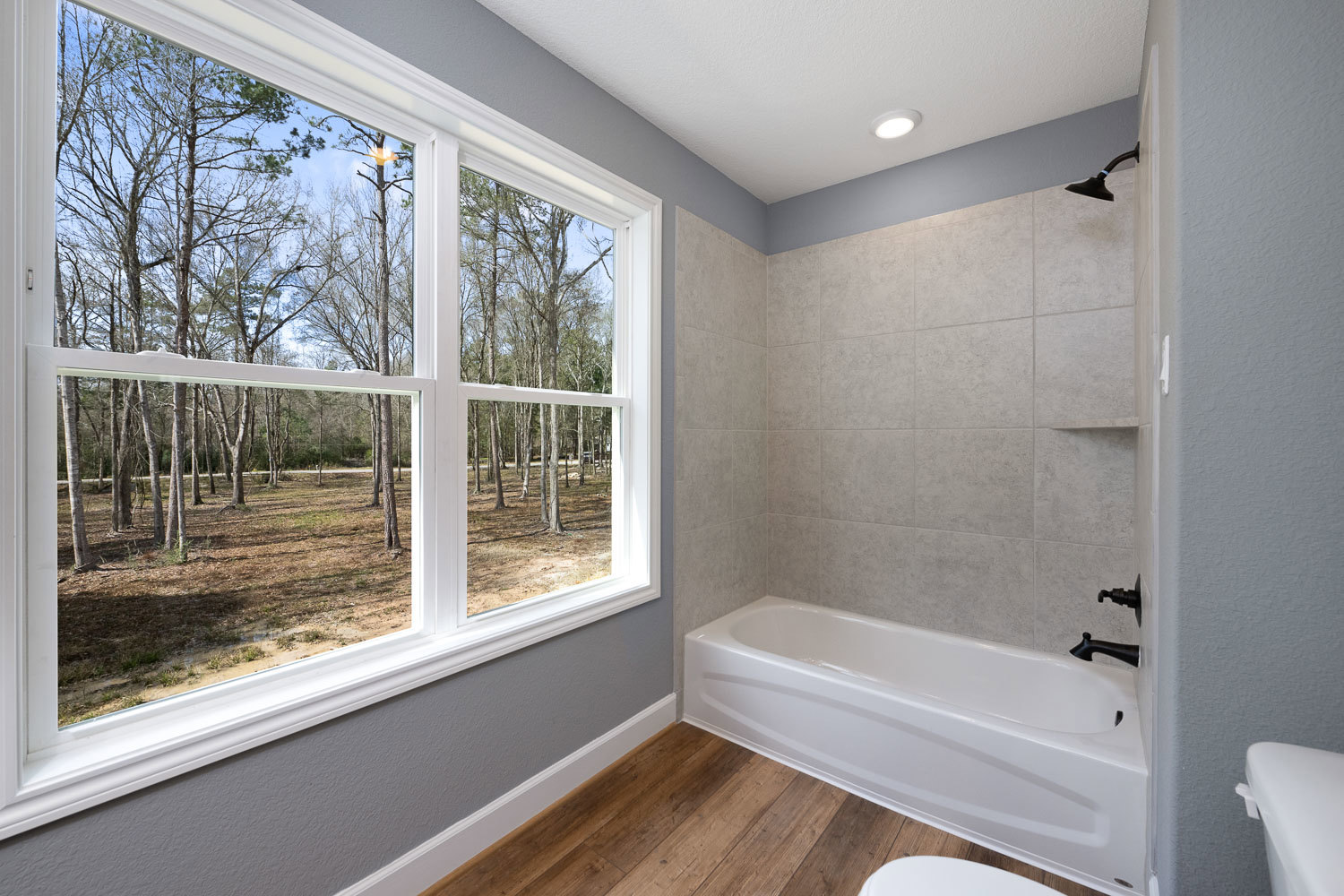 White freestanding bathtub beneath a large window with leafy trees visible outside, black metal faucet with round knob, light-colored walls, partial view of toilet in foreground