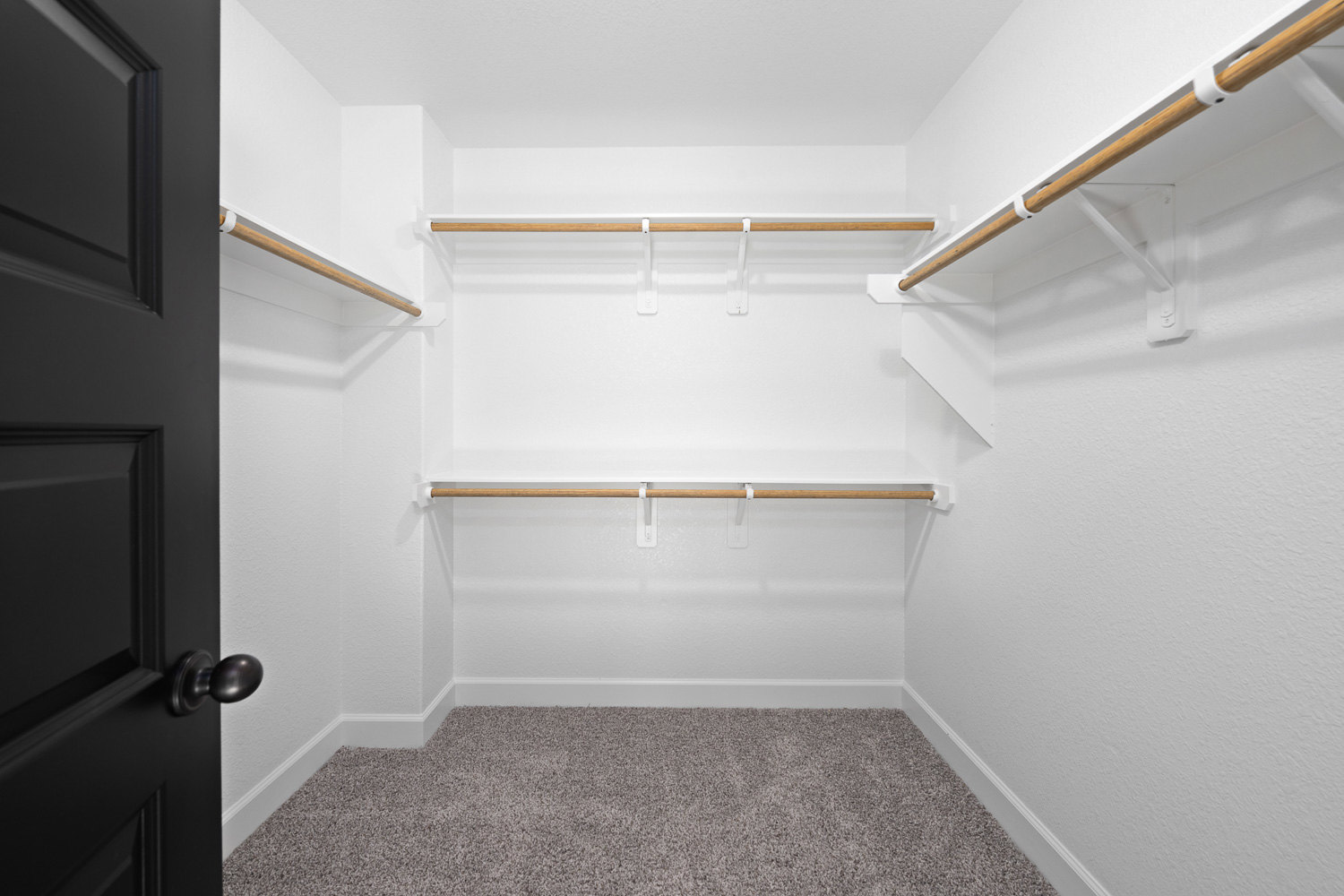 White closet with natural wood shelves, white walls, beige carpet flooring, black door with silver knob, and a white and wood hanging bar.