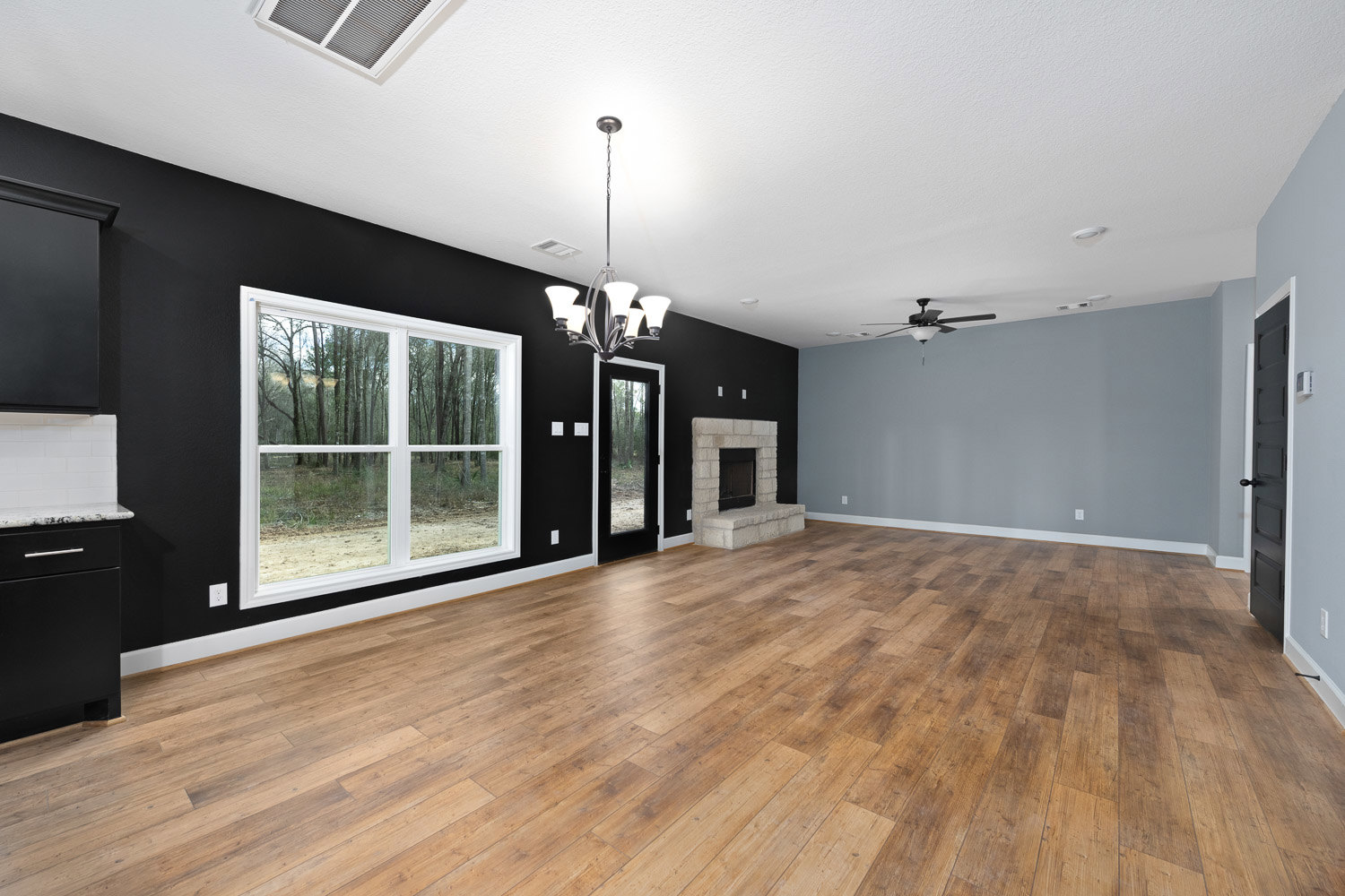 Living room with wide-plank hardwood flooring, modern black-framed fireplace, large window overlooking trees, white tile accent wall, and minimalist black hood.