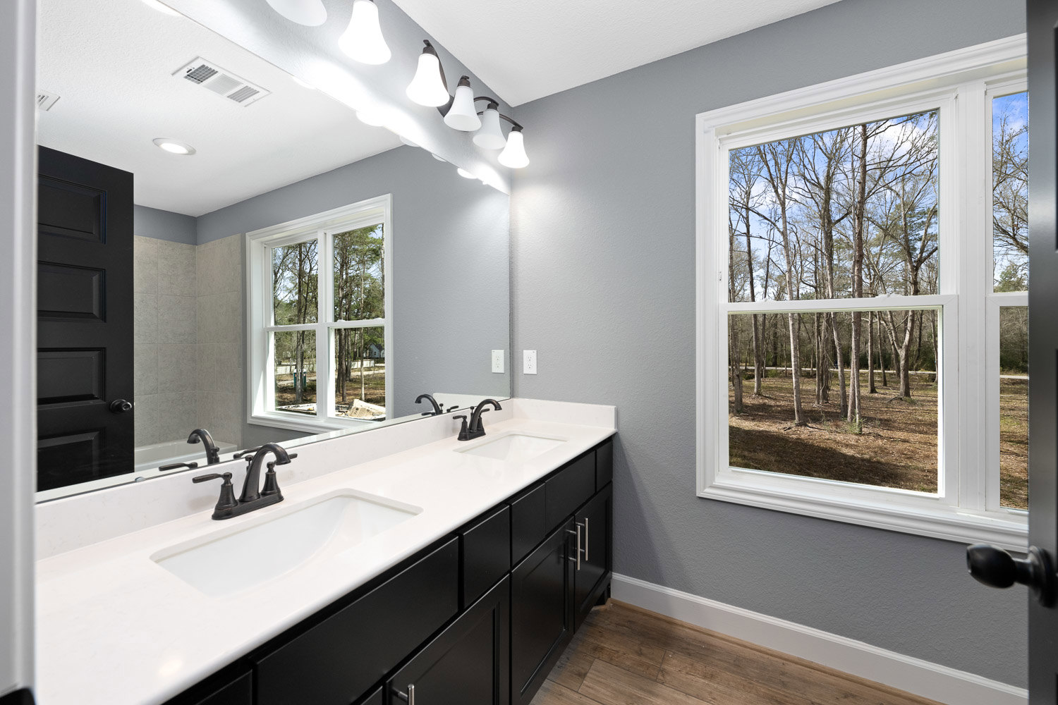 Modern bathroom featuring a rectangular window with forest view, white countertop sink, chrome faucet, large wall mirror, black door, and light gray tile walls.