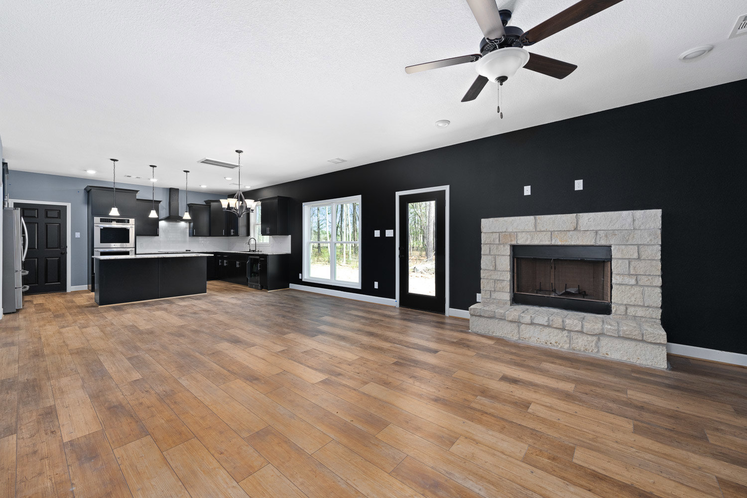 Living room with wood flooring, white ceiling, ceiling fan, black-framed fireplace, and large windows showing trees outside