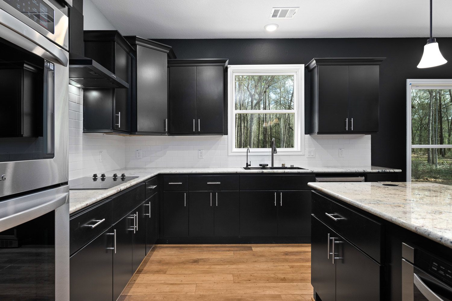 Black kitchen cabinets with silver handles, white countertops, stainless steel appliances, wood flooring, large window overlooking trees, modern ceiling light fixture, black accent