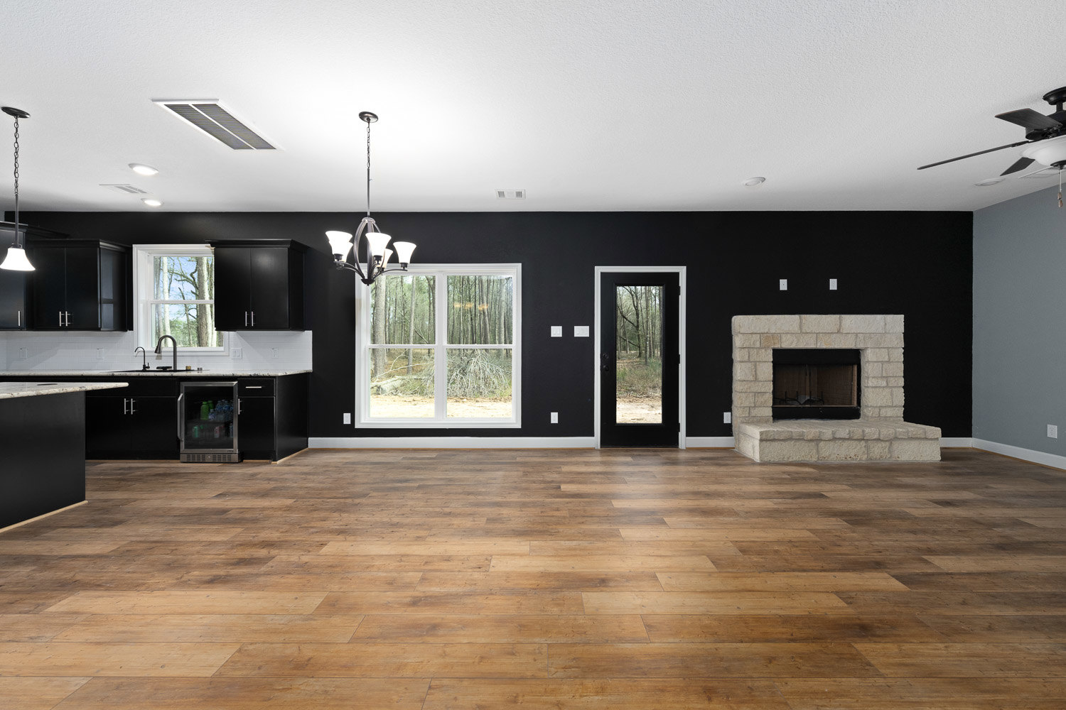 Living room with hardwood floors, black accent wall, modern fireplace, large window overlooking forested landscape, and glass-door refrigerator.