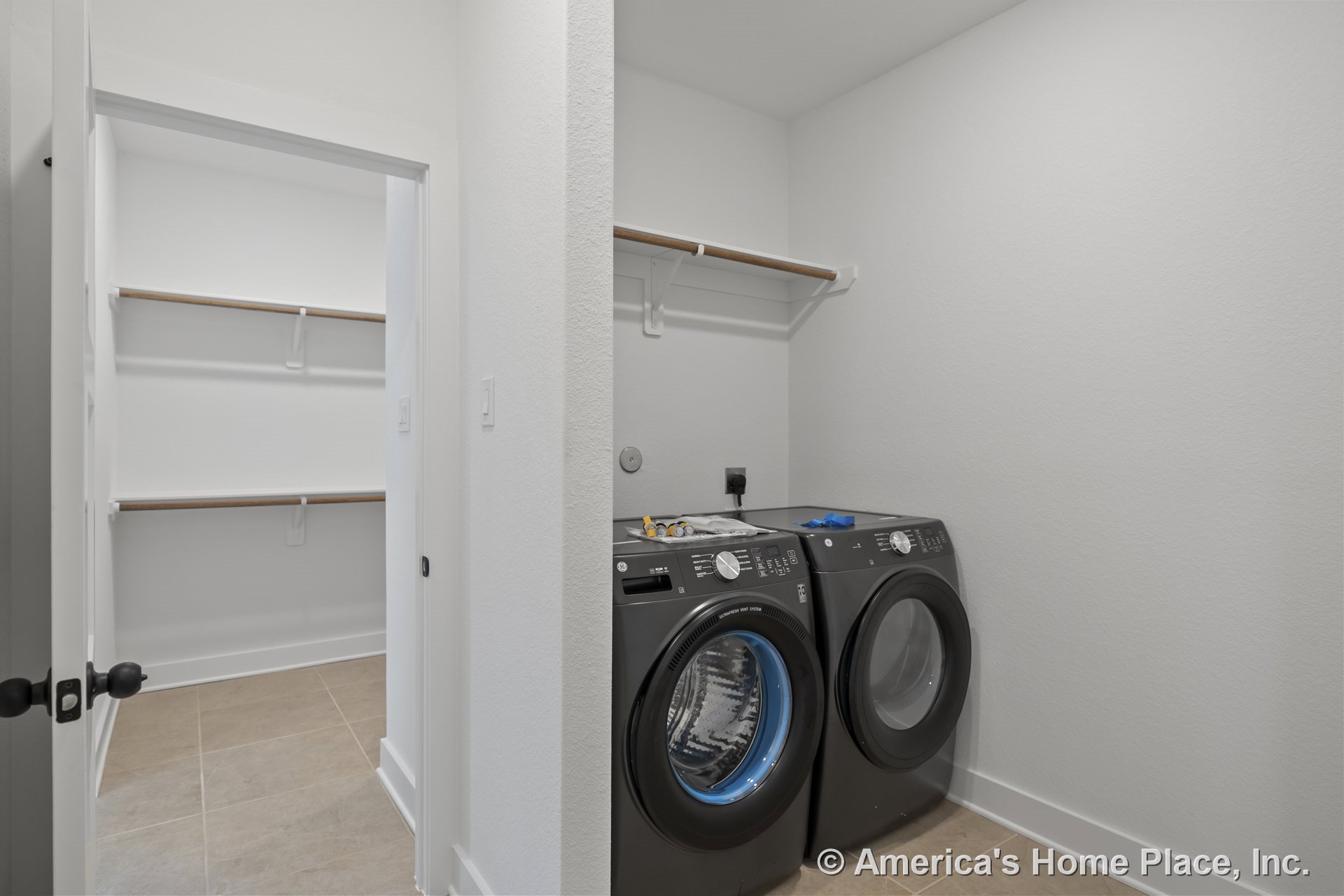 Front-loading washer and dryer set beside built-in wall shelving, tile flooring, white painted walls, baseboard trim, and adjacent walk-in closet with additional shelving.