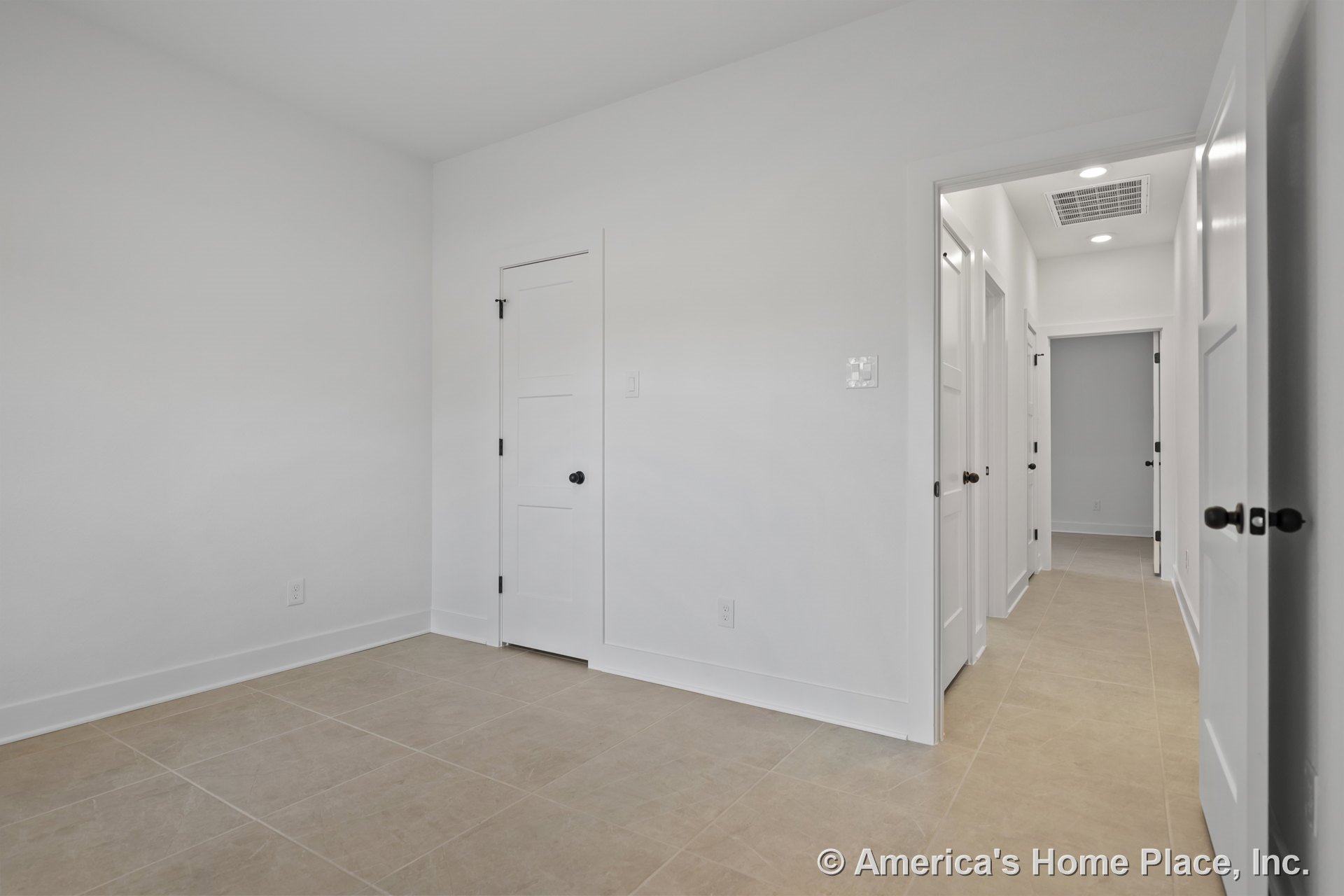 Wide hallway with large beige tile flooring, white paneled doors with black hardware, white walls, recessed ceiling lighting, and minimalist baseboards; adjacent room entry