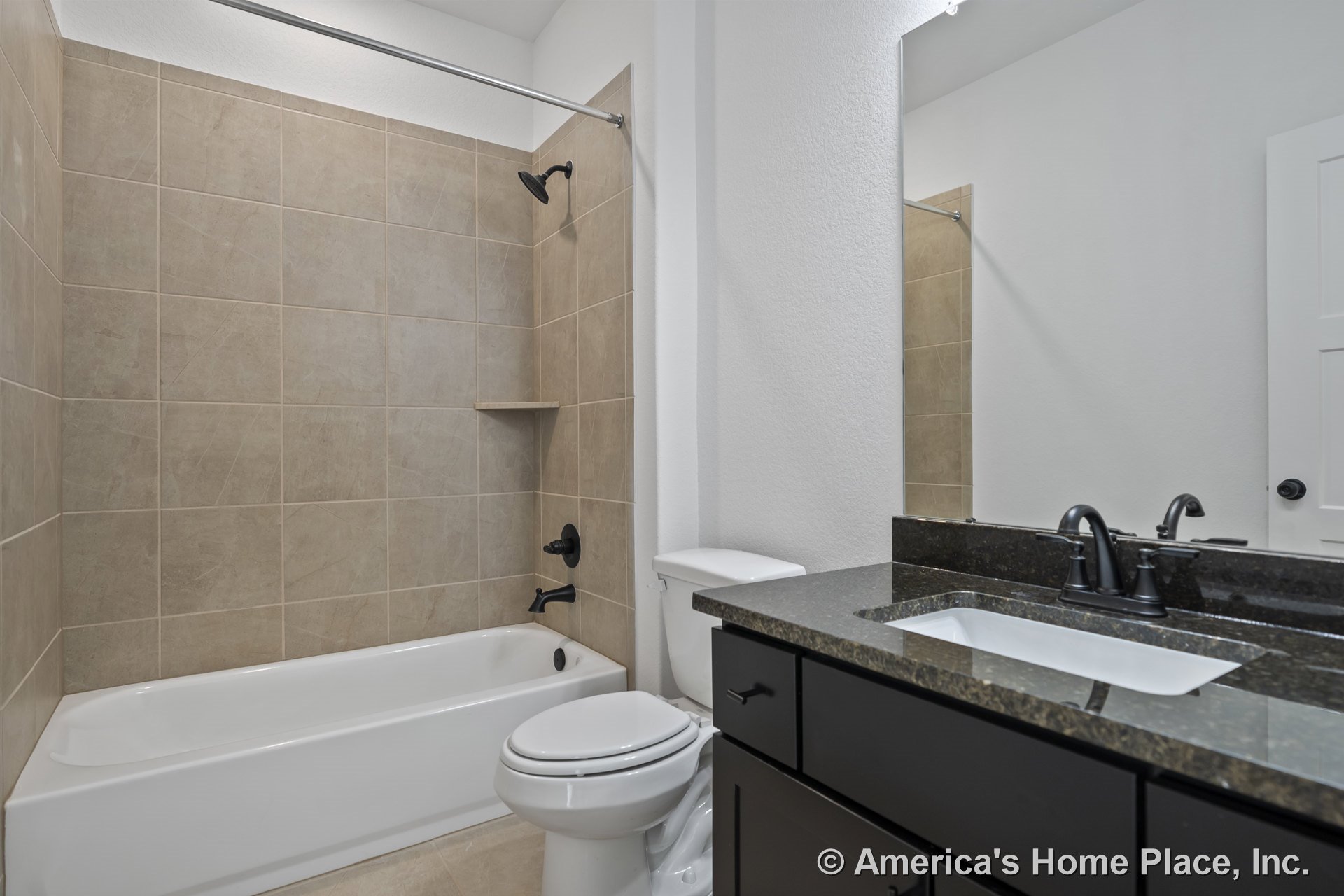Tan tile shower walls with white bathtub, black faucet fixtures, dark stone vanity countertop, large wall mirror, and black cabinet drawers.