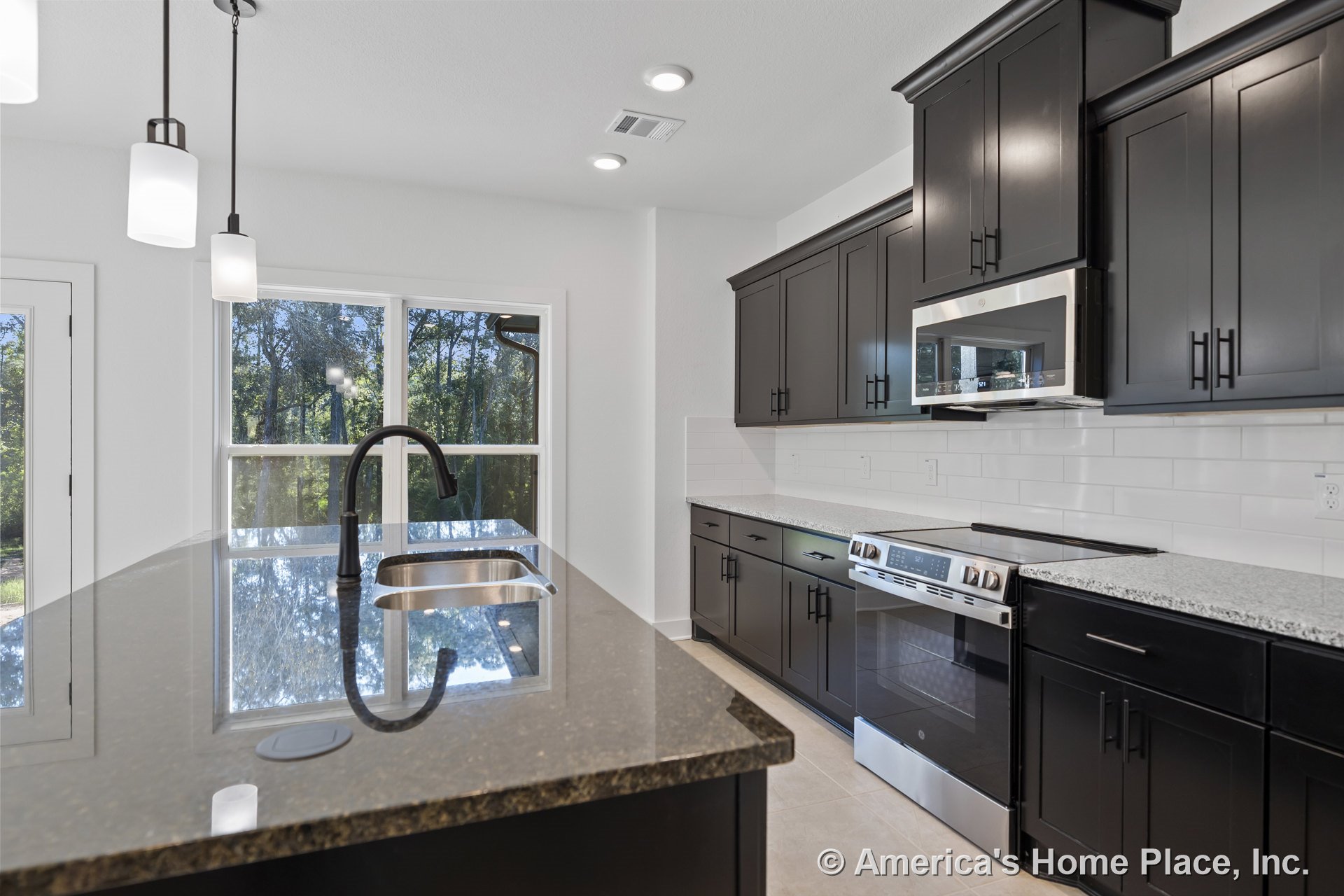 Spacious kitchen with large granite island featuring undermount sink, dark shaker cabinets, stainless steel range and built-in microwave, subway tile backsplash, pendant and