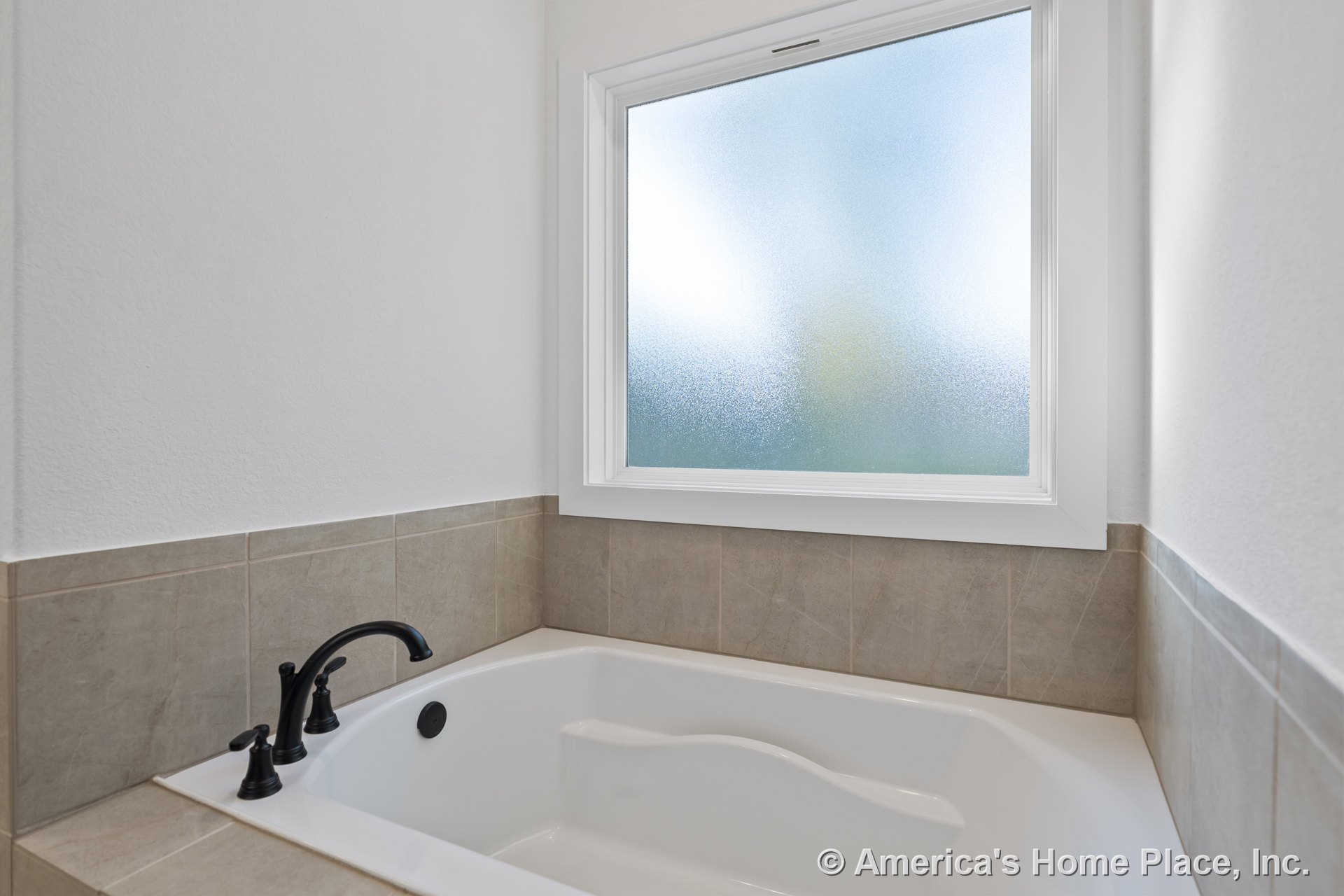 Built-in soaking tub with black faucet set in a recessed alcove, surrounded by beige tile; frosted glass window with white trim above tub; neutral wall paint and modern permanent