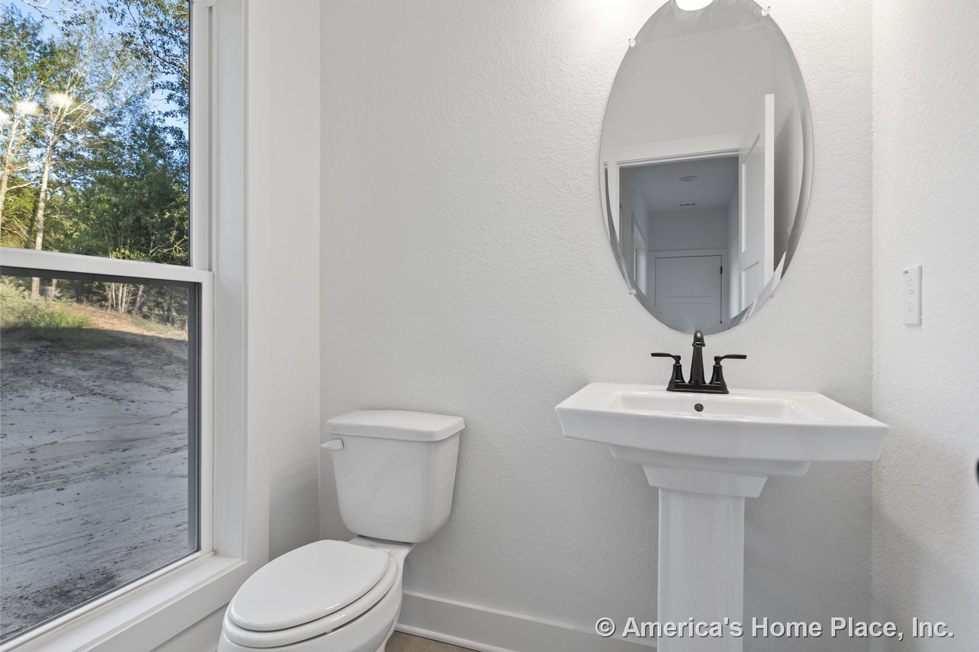 Pedestal sink with black faucet beneath oval wall-mounted mirror, large window with white trim, white toilet, and light-colored flooring in a minimalist bathroom.