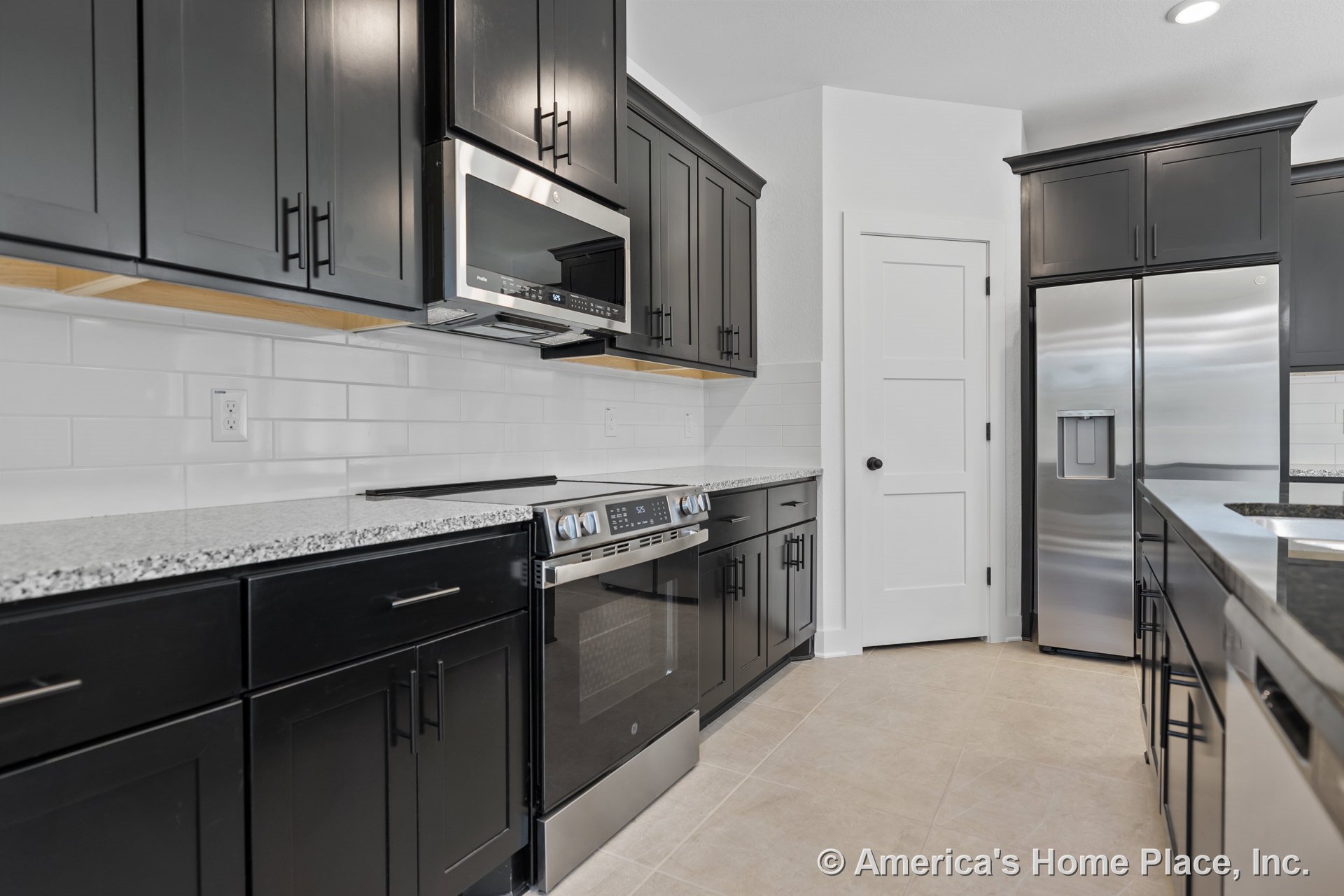 Black shaker cabinets with granite countertops, stainless steel refrigerator, built-in microwave, subway tile backsplash, and tile flooring in a modern kitchen.