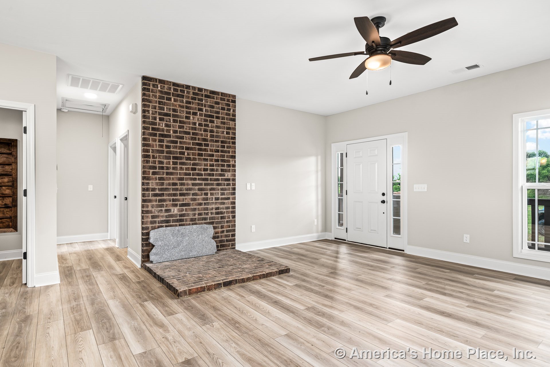 Brick accent wall with hearth, light wood plank flooring, white trim and baseboards, ceiling fan with light, front door featuring sidelights, large windows, and neutral wall paint