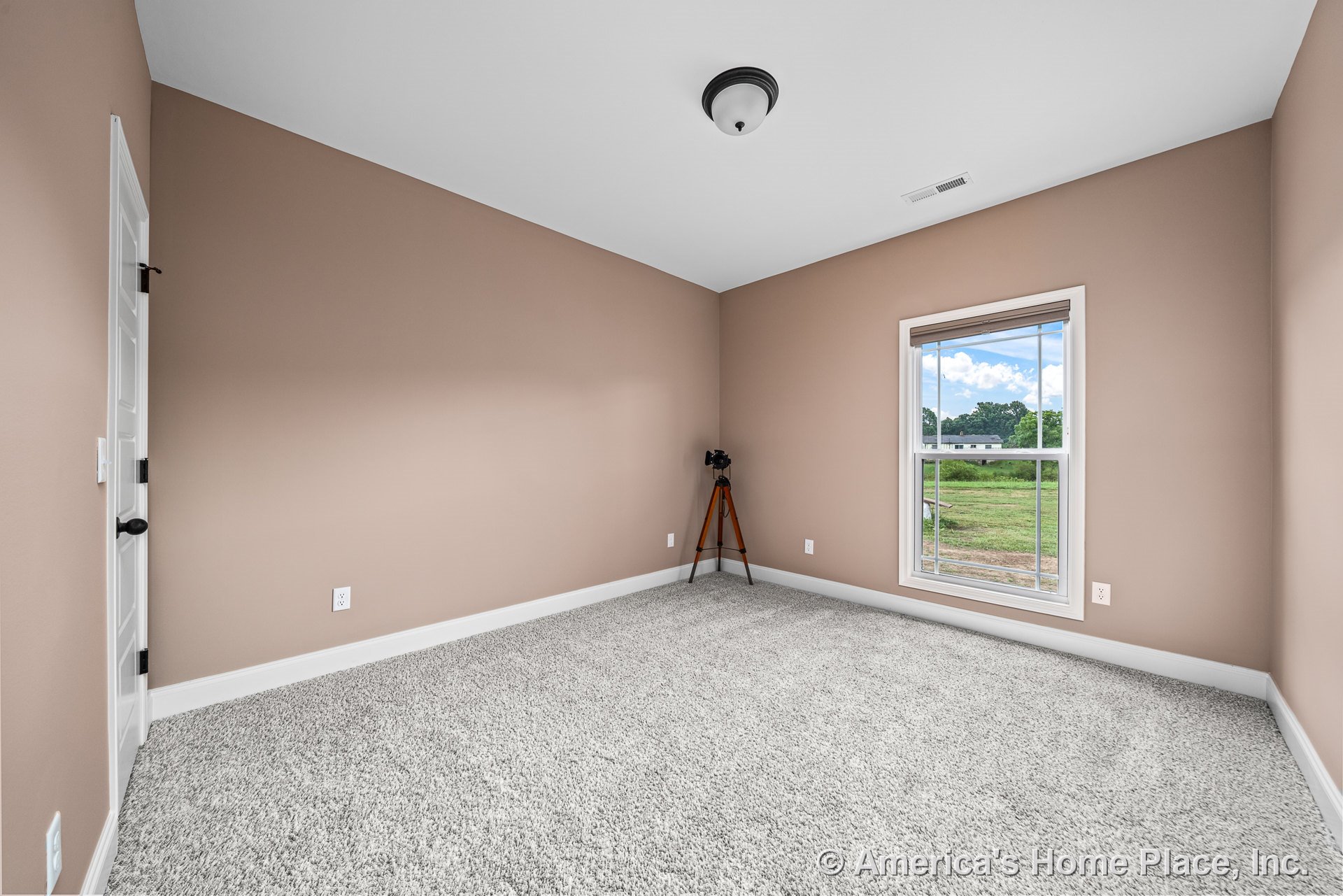 Beige painted walls with white baseboards and trim, single double-hung window, light gray carpet flooring, white paneled door, and flush mount ceiling light in a modern bedroom.
