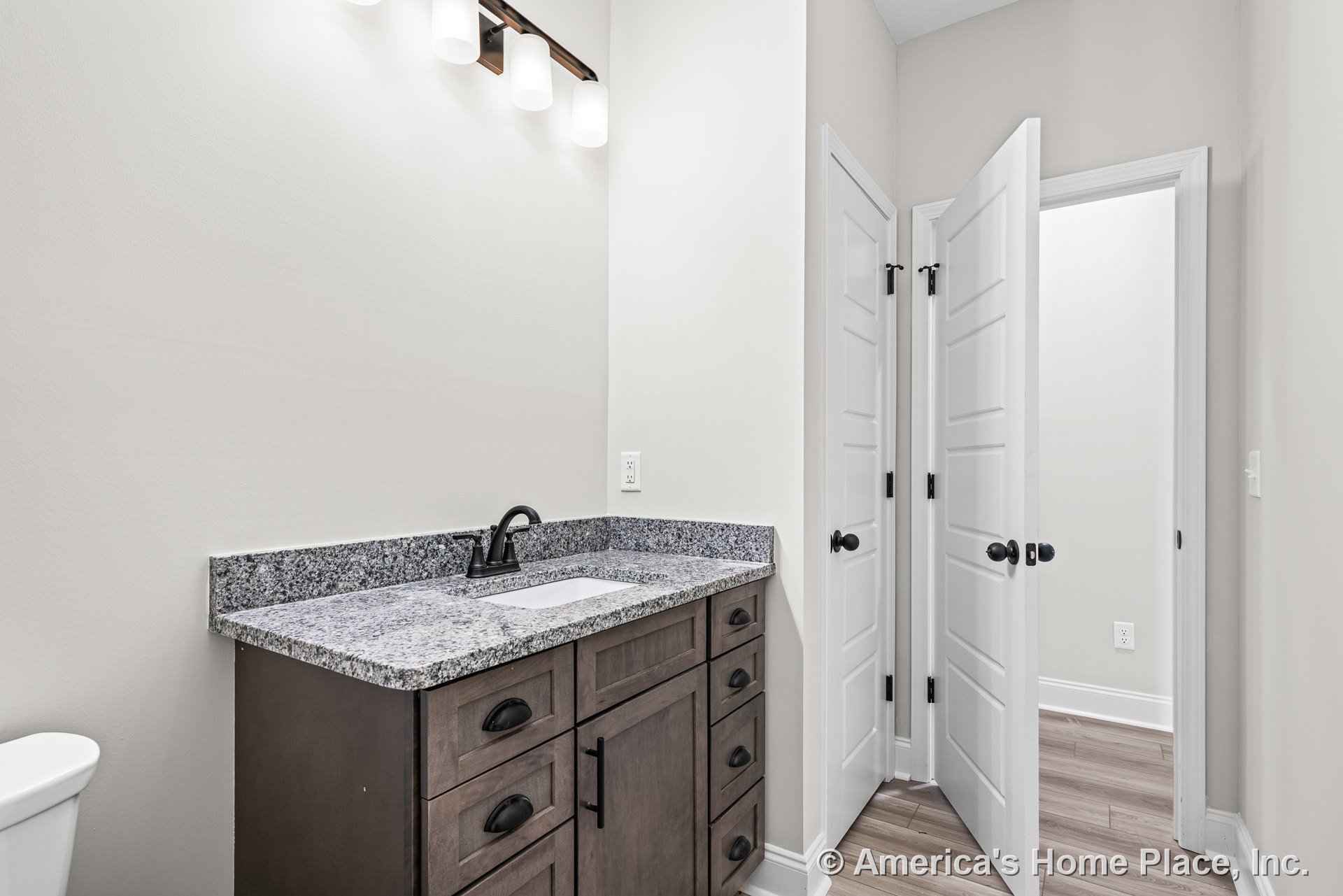 Granite vanity countertop with dark wood cabinetry, black faucet and hardware, white paneled doors, overhead built-in vanity lighting, wood-look tile flooring, and neutral wall