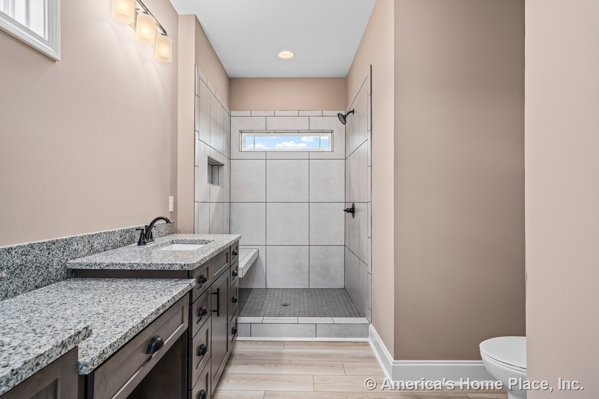 Walk-in shower with large tiled walls, built-in bench, horizontal window, granite double vanity with dark wood cabinetry, wall-mounted lighting fixtures, and modern plumbing