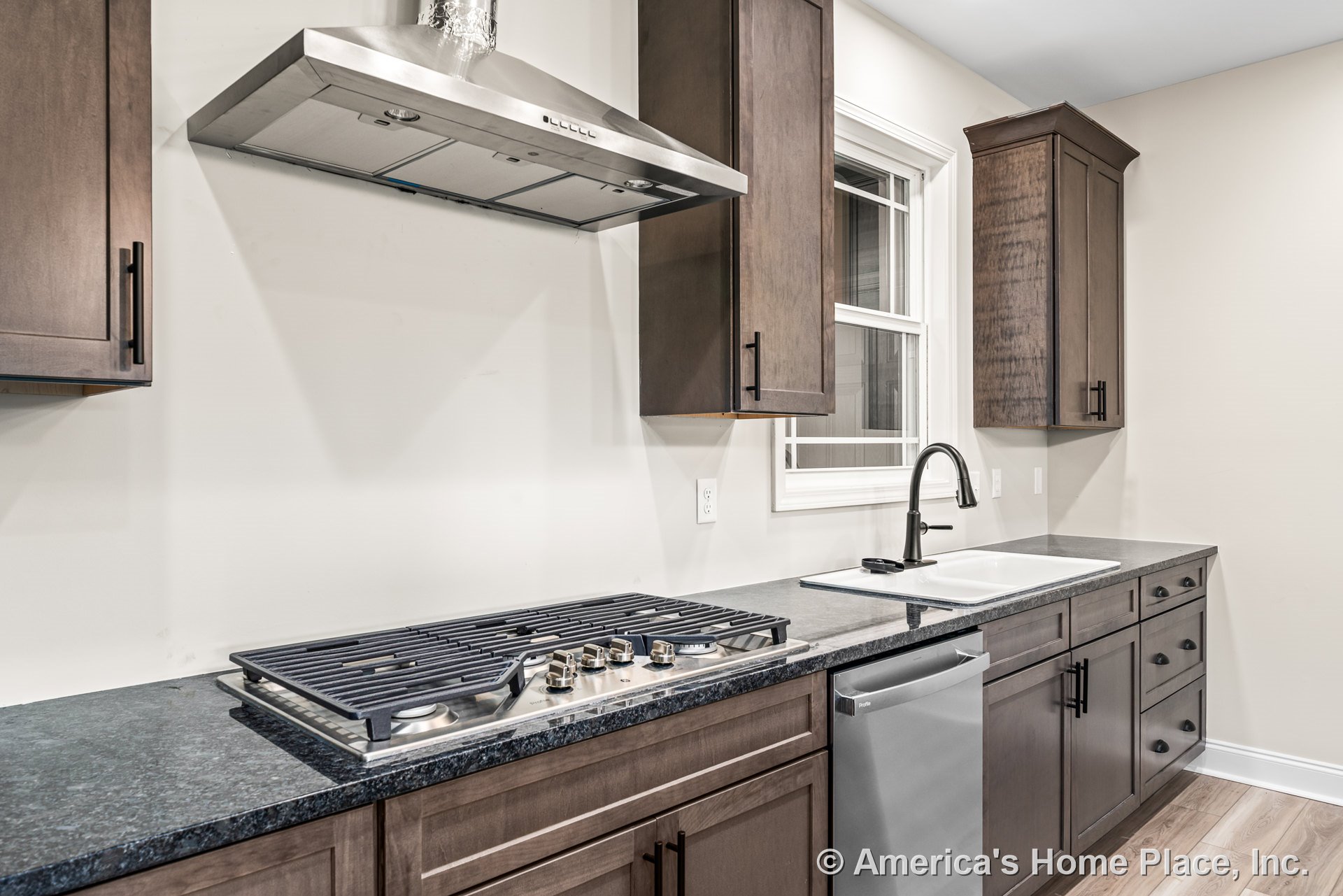 Dark wood cabinetry with stainless steel range hood and built-in gas cooktop, black stone countertops, undermount sink with black faucet beneath window, integrated dishwasher