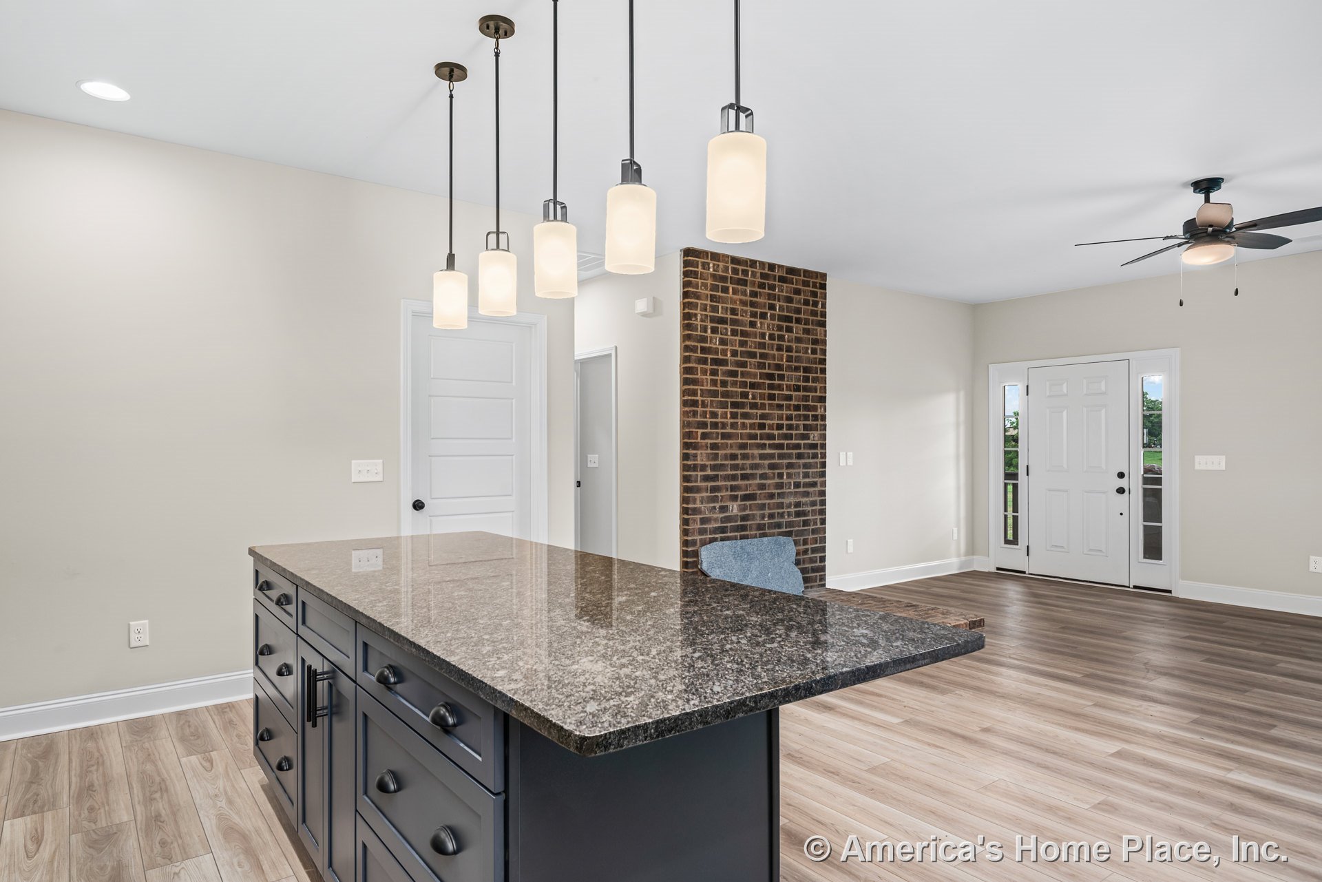 Spacious kitchen with a large granite island and dark shaker cabinets, open to a living area featuring a brick accent wall, light wood plank flooring, pendant lights, white paneled