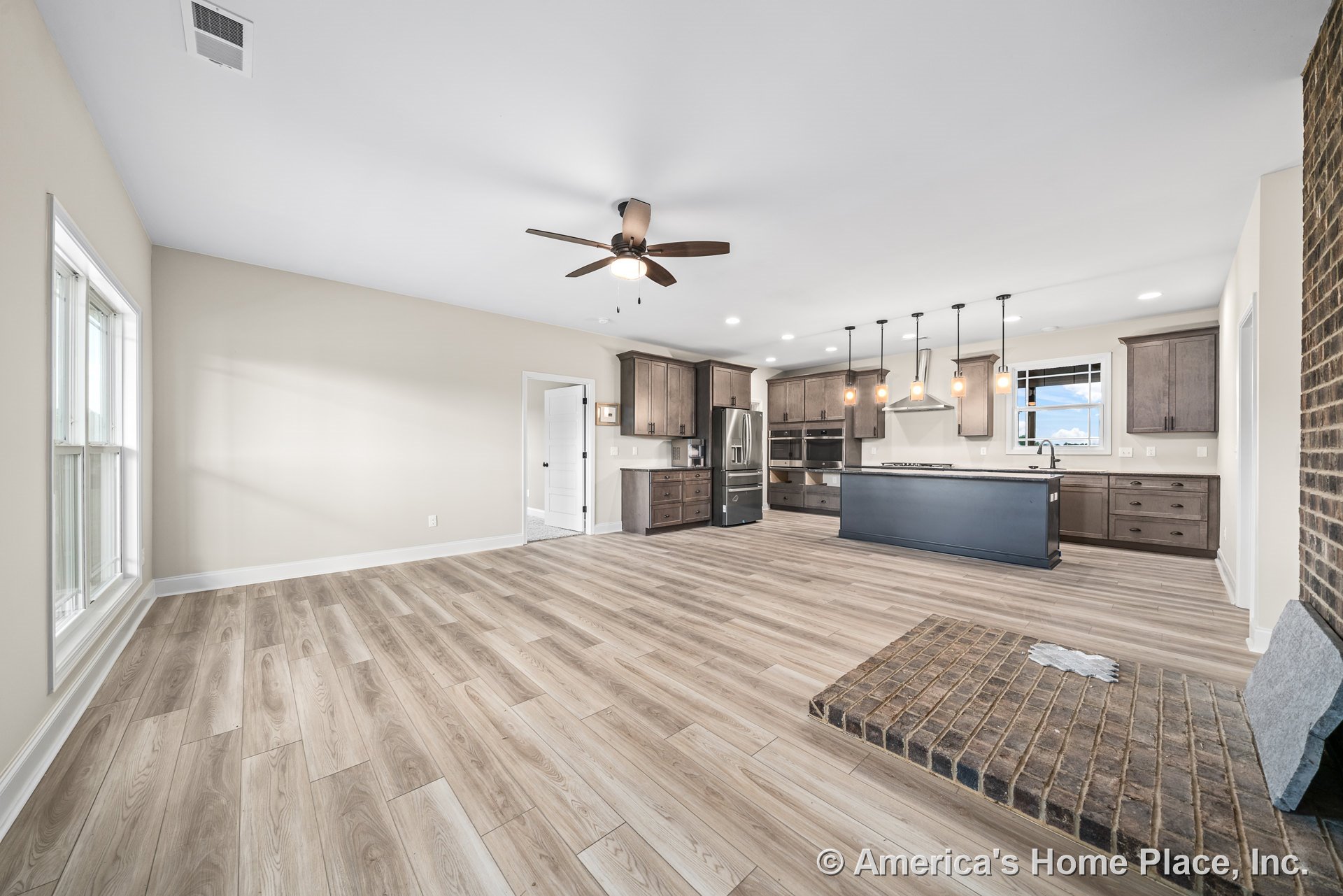 Open concept living area with wood-look plank flooring, brick fireplace hearth, kitchen island with dark cabinetry and stainless steel appliances, pendant lights above island