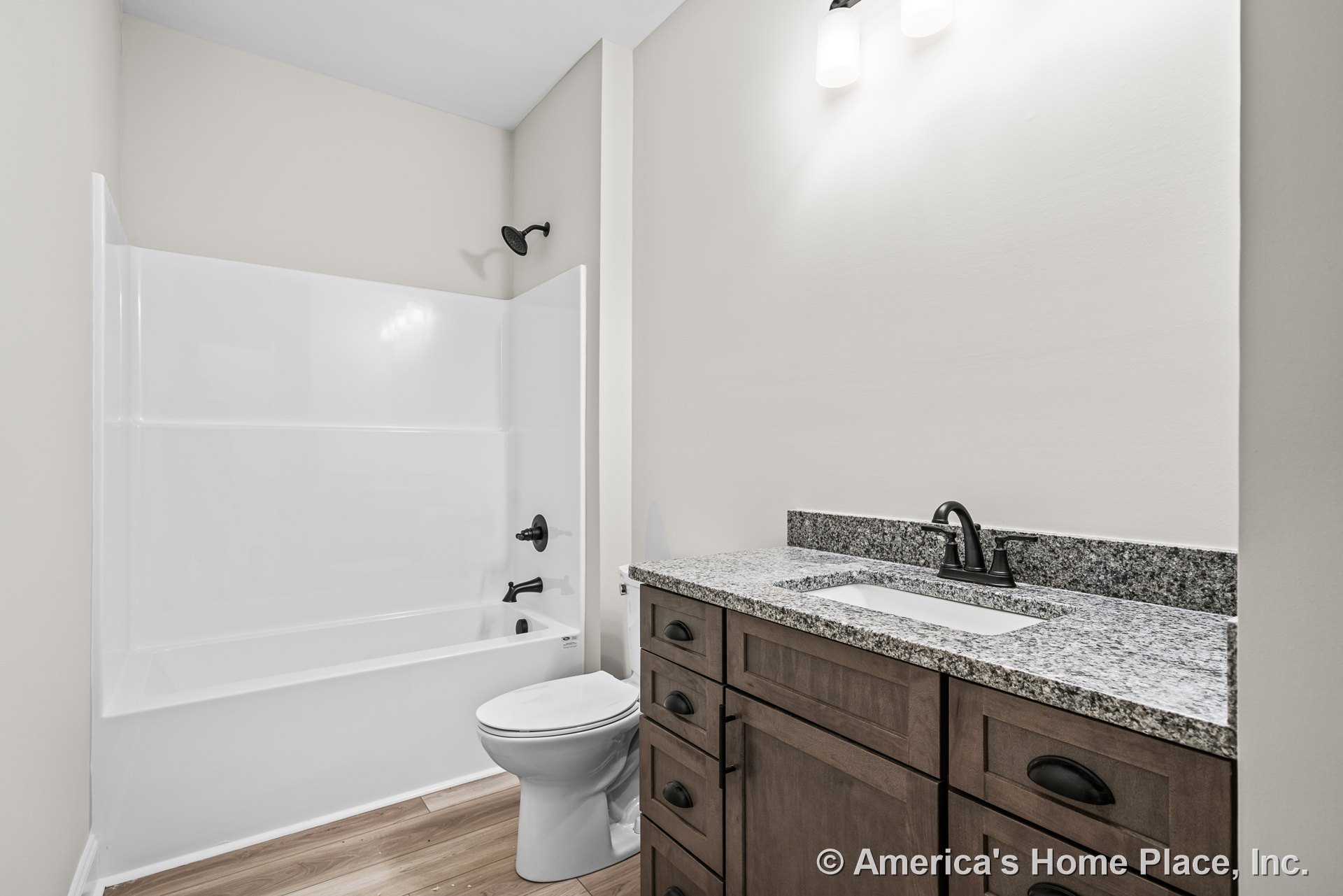 Wood vanity with granite countertop, built-in sink, matte black faucet and hardware, white tub-shower unit, overhead vanity lighting, light wood flooring, and neutral walls.