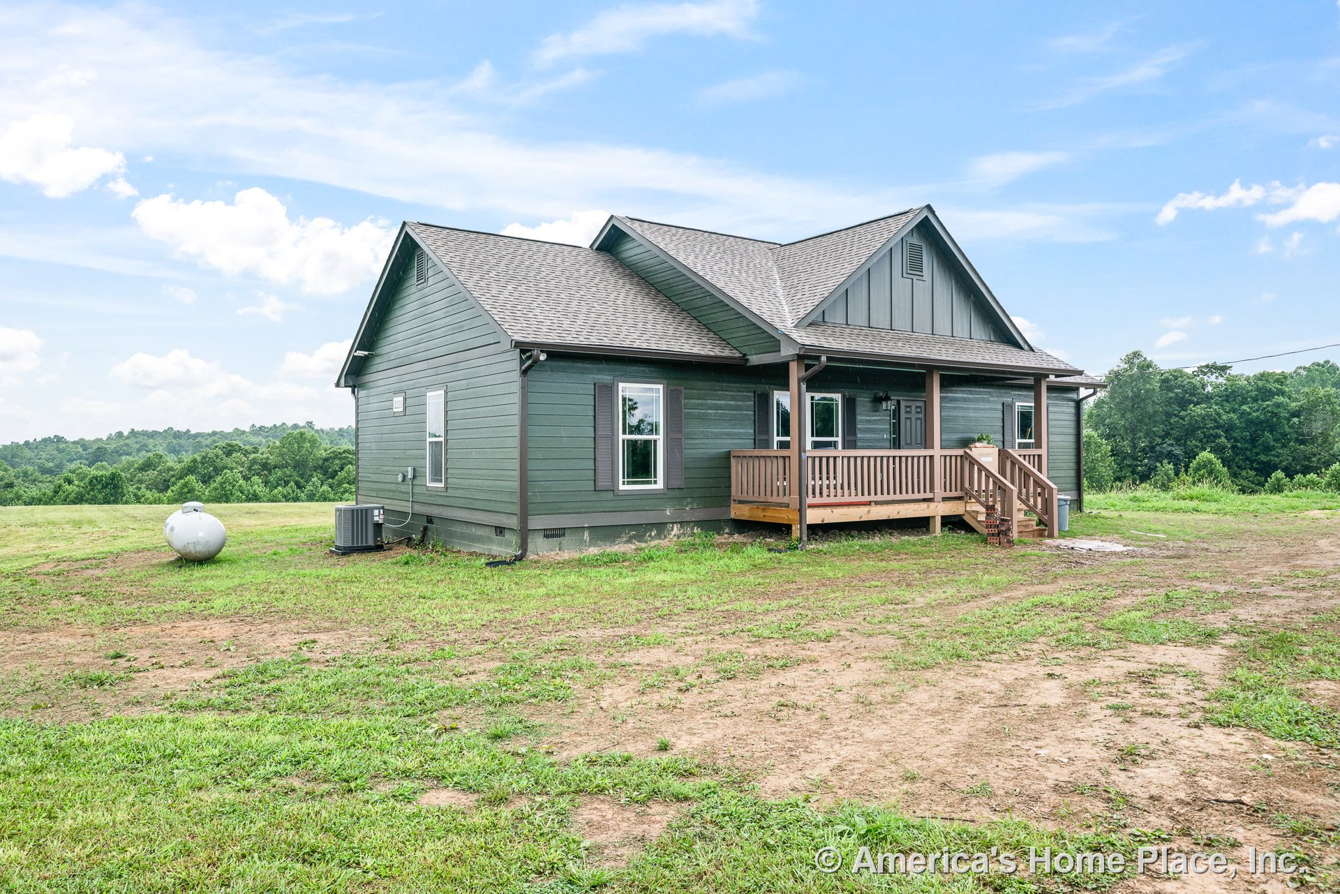 Covered front porch with wood railings and steps, green horizontal siding, gabled roof with asphalt shingles, multiple double-hung windows, wood posts supporting the porch