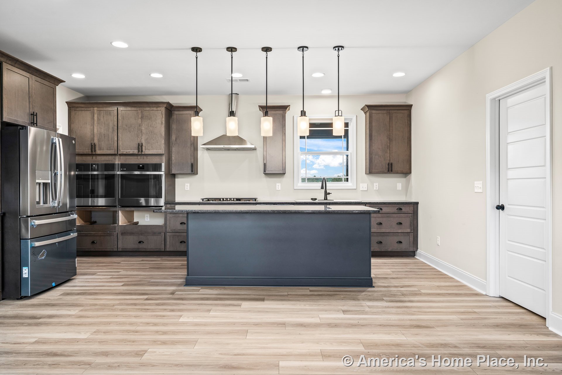 Shaker-style wood cabinetry with stainless steel appliances, central kitchen island featuring dark countertops, pendant lighting above, light wood flooring, window above sink