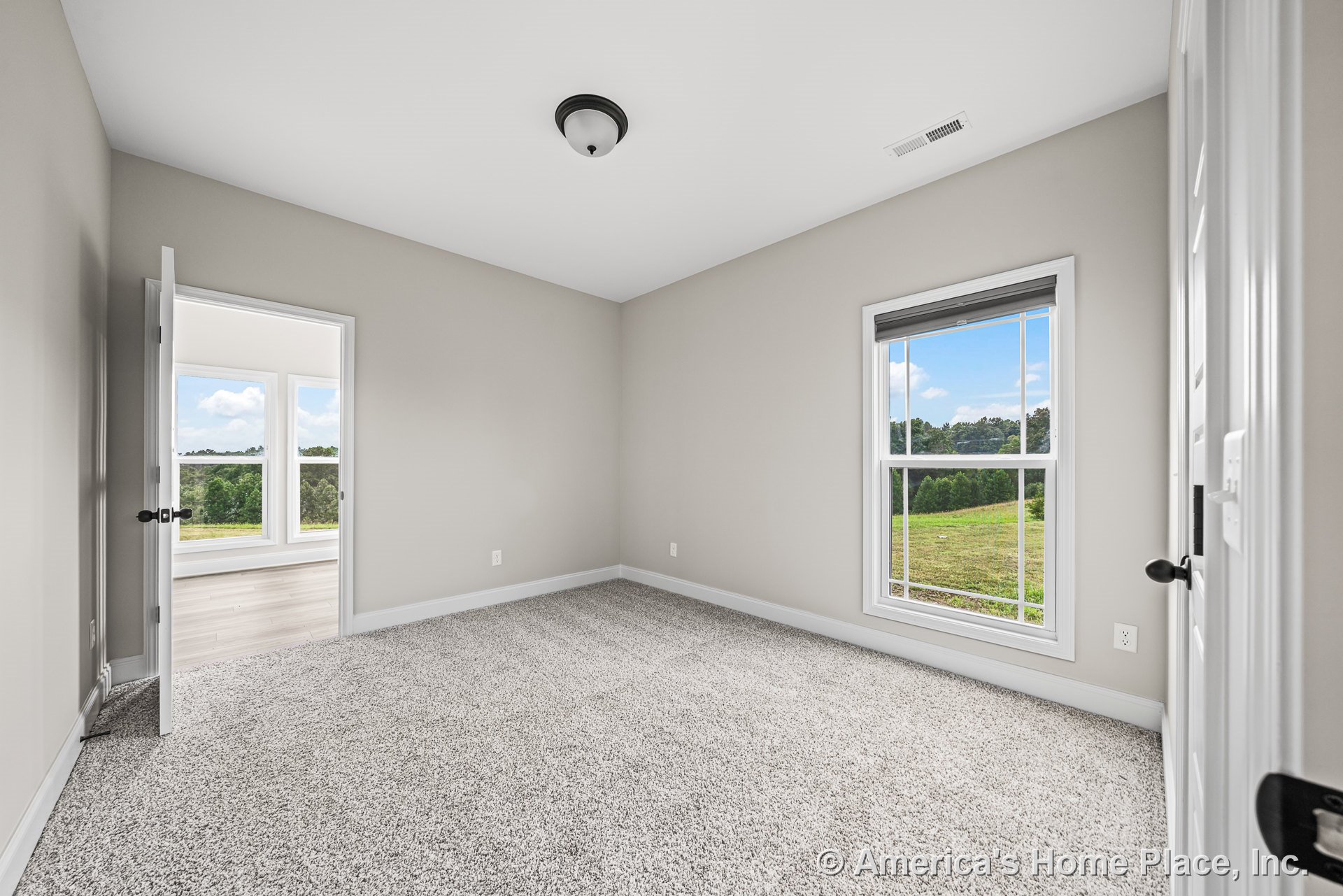 Bedroom with light gray carpet flooring, neutral painted walls, white baseboards, large window with white trim, flush mount ceiling light, and doorway leading to adjacent room with