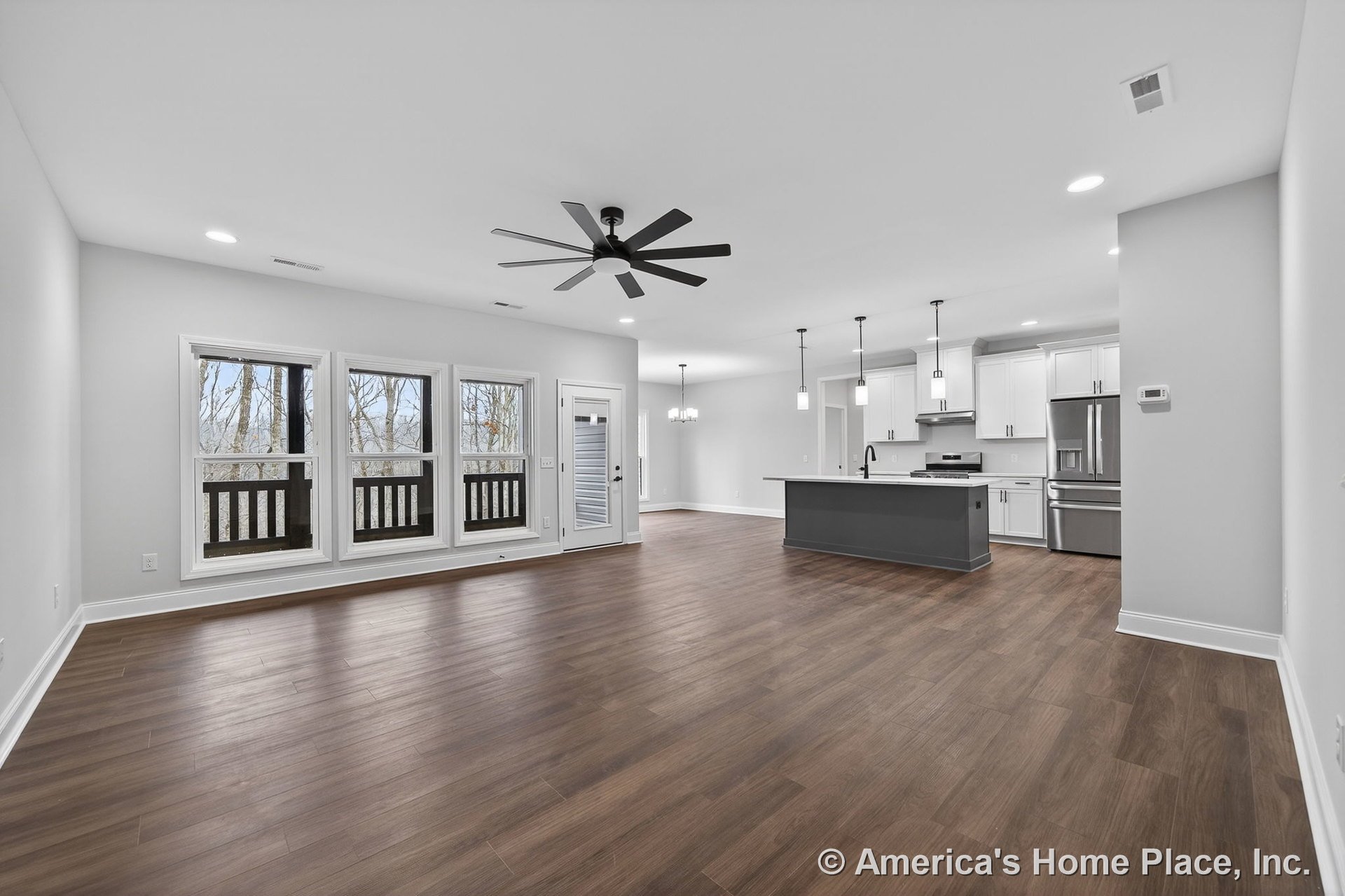 Open-concept living area with wide plank wood flooring, large windows with trim, modern ceiling fan, kitchen island with pendant lights, white shaker cabinets, stainless steel