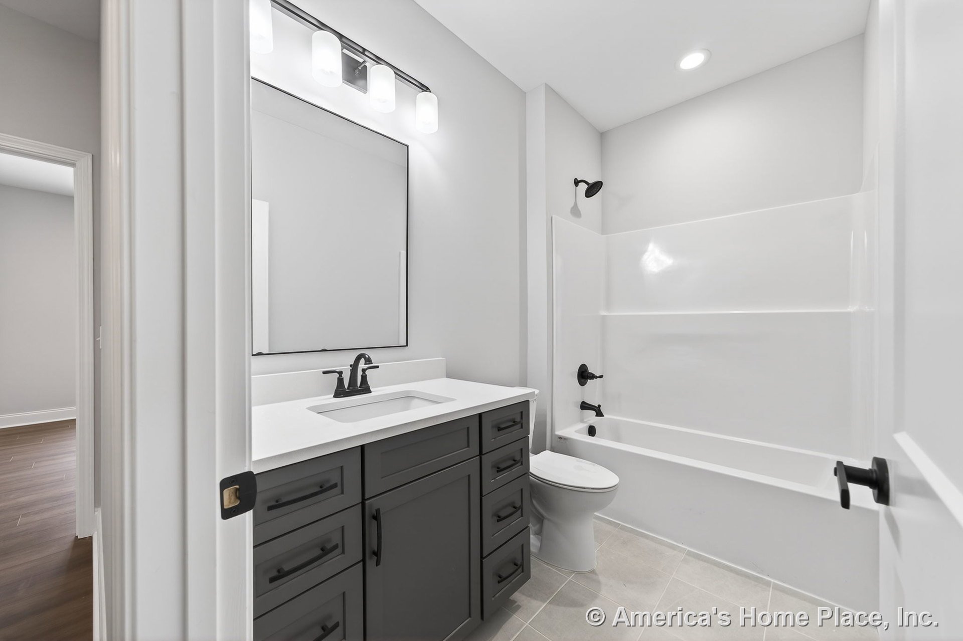Dark vanity with matte black hardware and white solid surface countertop, integrated bathtub and shower with neutral wall color, large framed mirror above three-light vanity