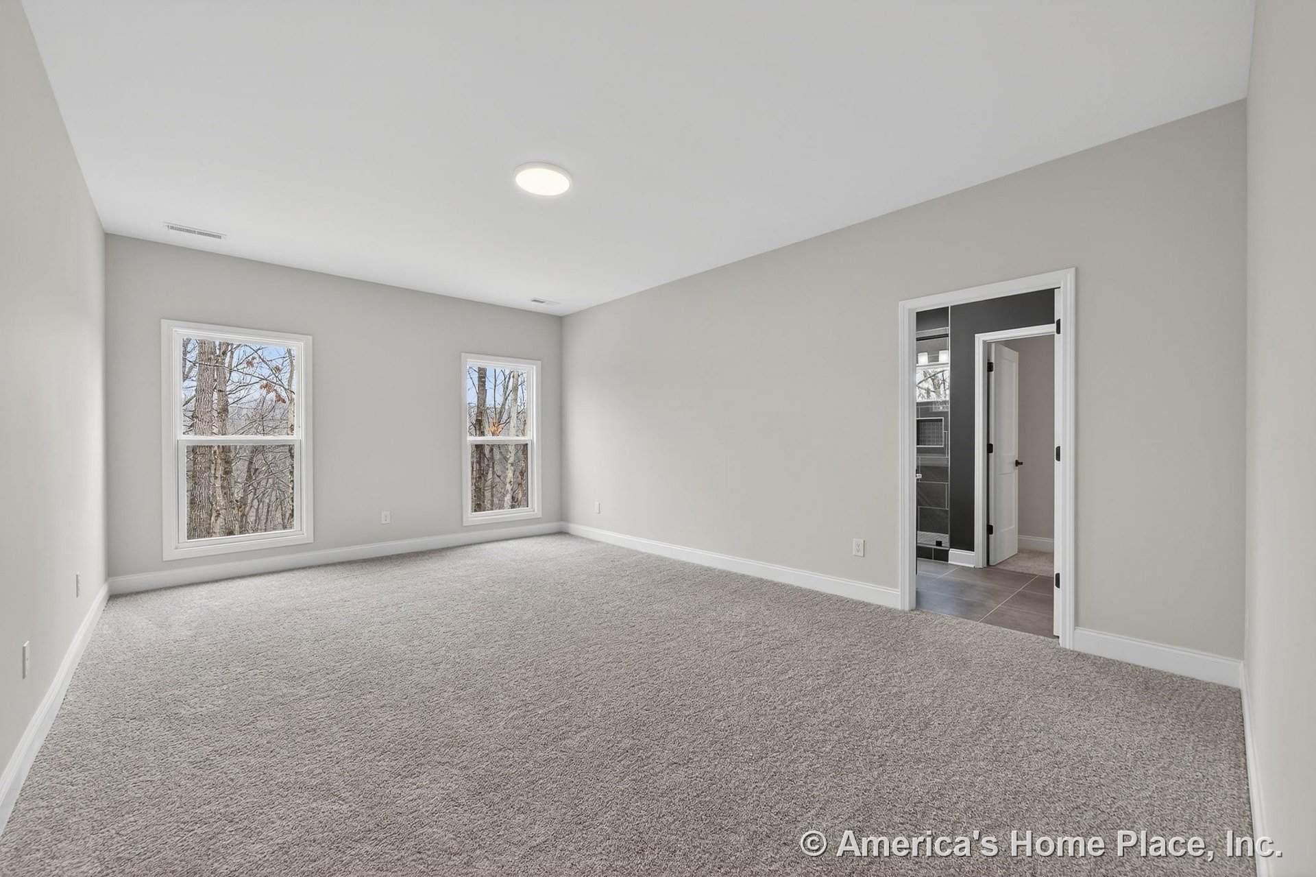 Spacious bedroom with carpeted floor, two large windows allowing natural light, neutral wall paint, white baseboards, recessed ceiling light, and doorway leading to bathroom.