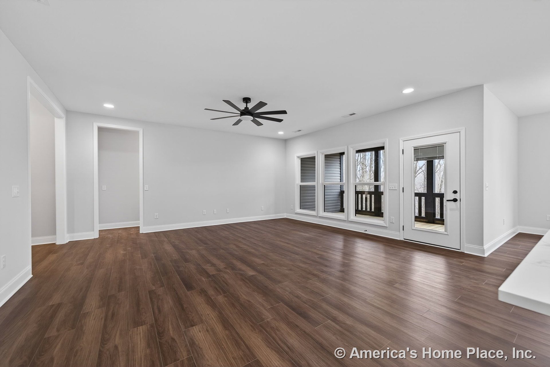 Open living room with wide plank wood-look flooring, expansive window wall, white trim, ceiling fan, recessed lighting, glass door leading to porch, and abundant natural light.