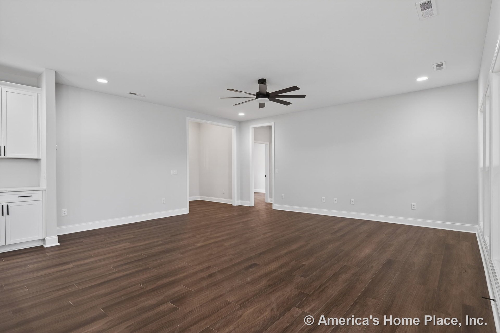 Open living room with wide dark wood-look flooring, white baseboards, recessed ceiling lights, large modern ceiling fan, built-in cabinetry, neutral walls, multiple windows, and
