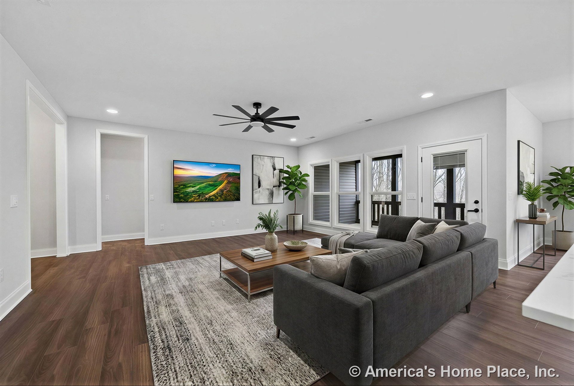 Wide plank wood floors, recessed ceiling lights, ceiling fan, large windows with trim, glass exterior door, wall-mounted television, white painted walls, baseboards, and open