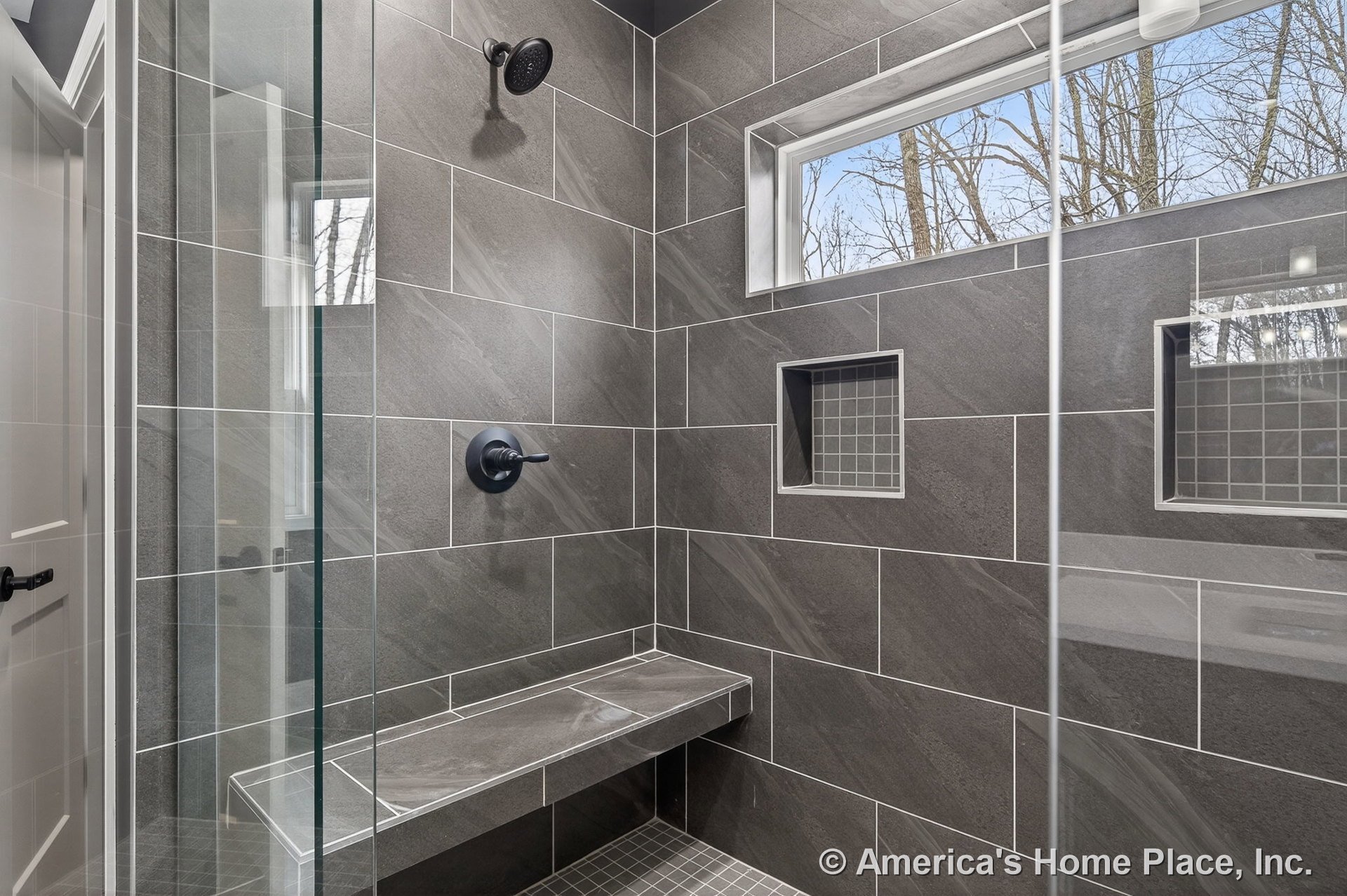 Walk-in shower with large gray tile walls, built-in bench, two recessed niches, matte black fixtures, glass door, and horizontal window providing natural light.