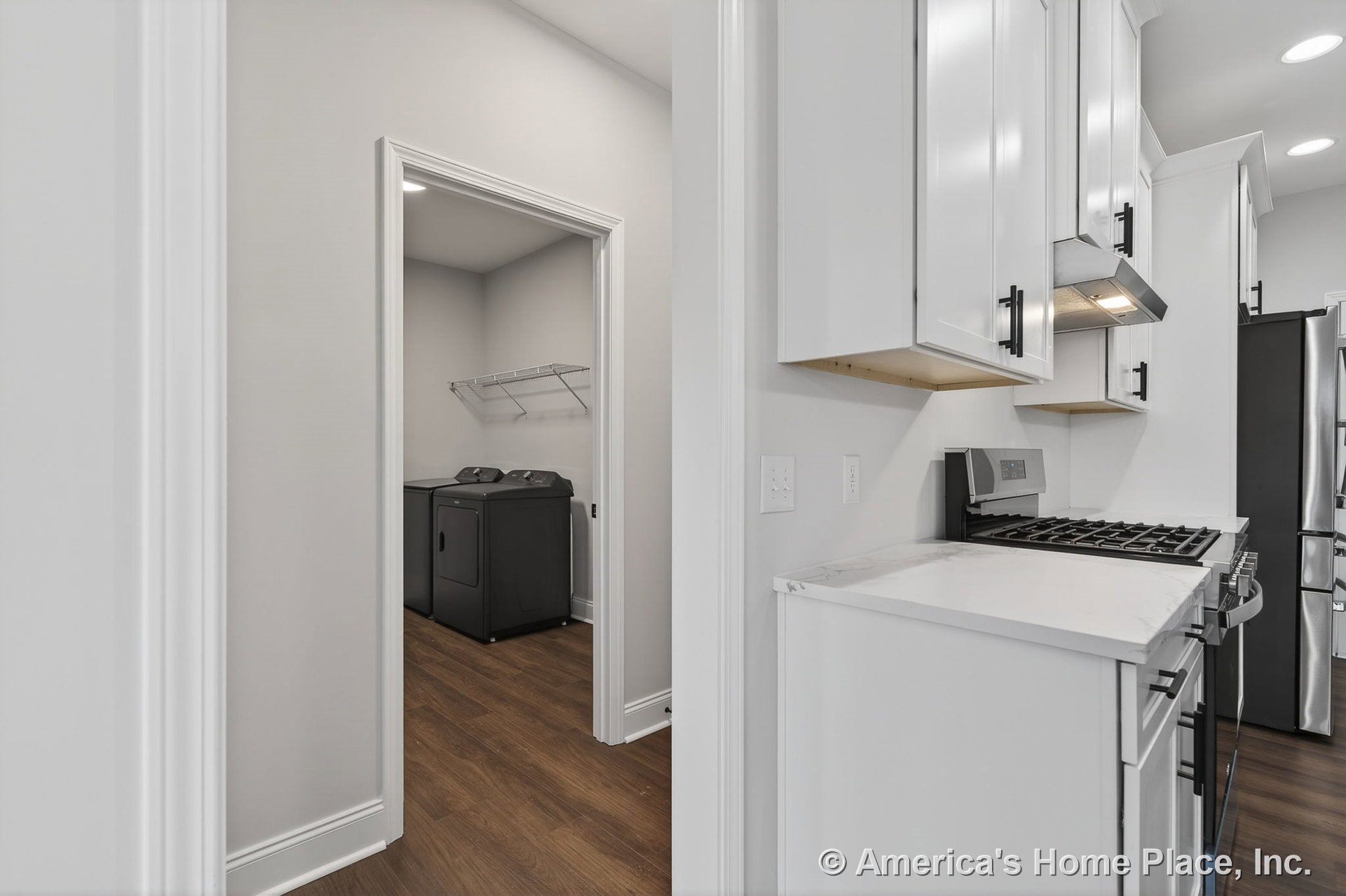 White kitchen cabinetry and marble-look countertops with stainless steel range hood and gas stove, open to laundry room featuring wood plank flooring, washer and dryer, wire