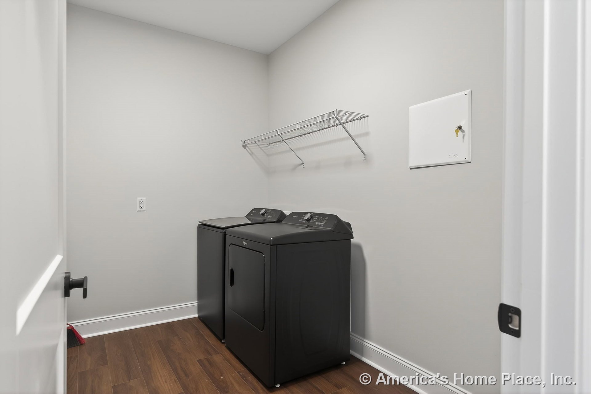 Black washer and dryer units beneath a wire wall shelf, wood-look flooring, white baseboard trim, neutral painted walls, and flush ceiling in a laundry room.