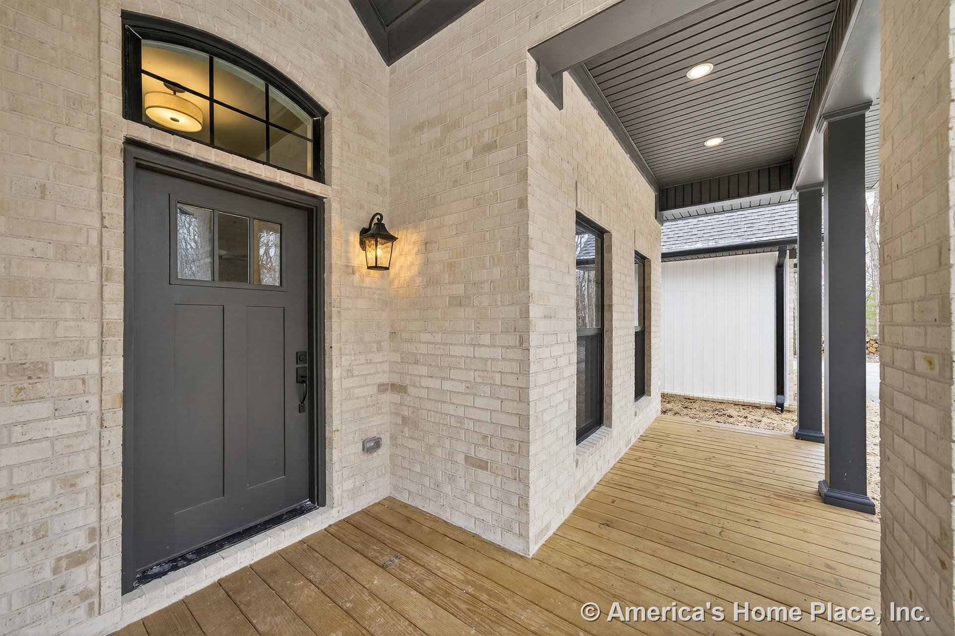 Covered front porch with light brick walls, dark gray front door topped by a transom window, wood plank flooring, black framed windows, paneled dark ceiling with recessed lights