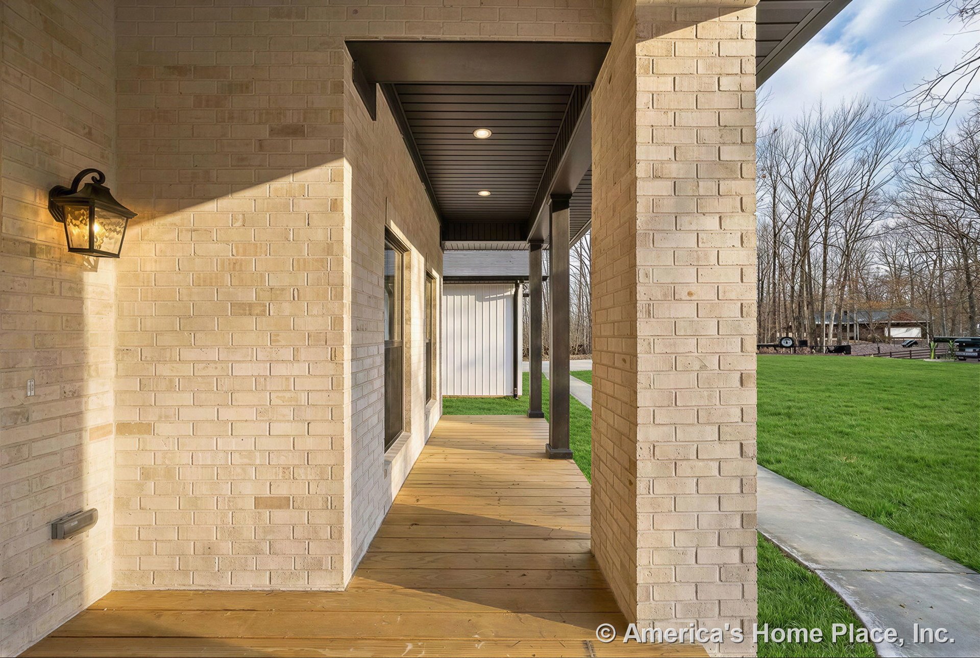 Covered porch with light brick columns, wood flooring, recessed ceiling lights, black metal trim, large glass window, and wall-mounted lantern.