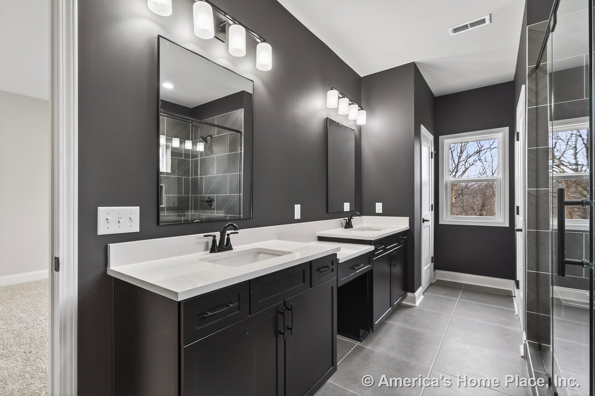 Dual vanity bathroom with black shaker cabinets, white solid surface countertops, large gray tile flooring, frameless glass shower enclosure, modern vanity lighting, matte black