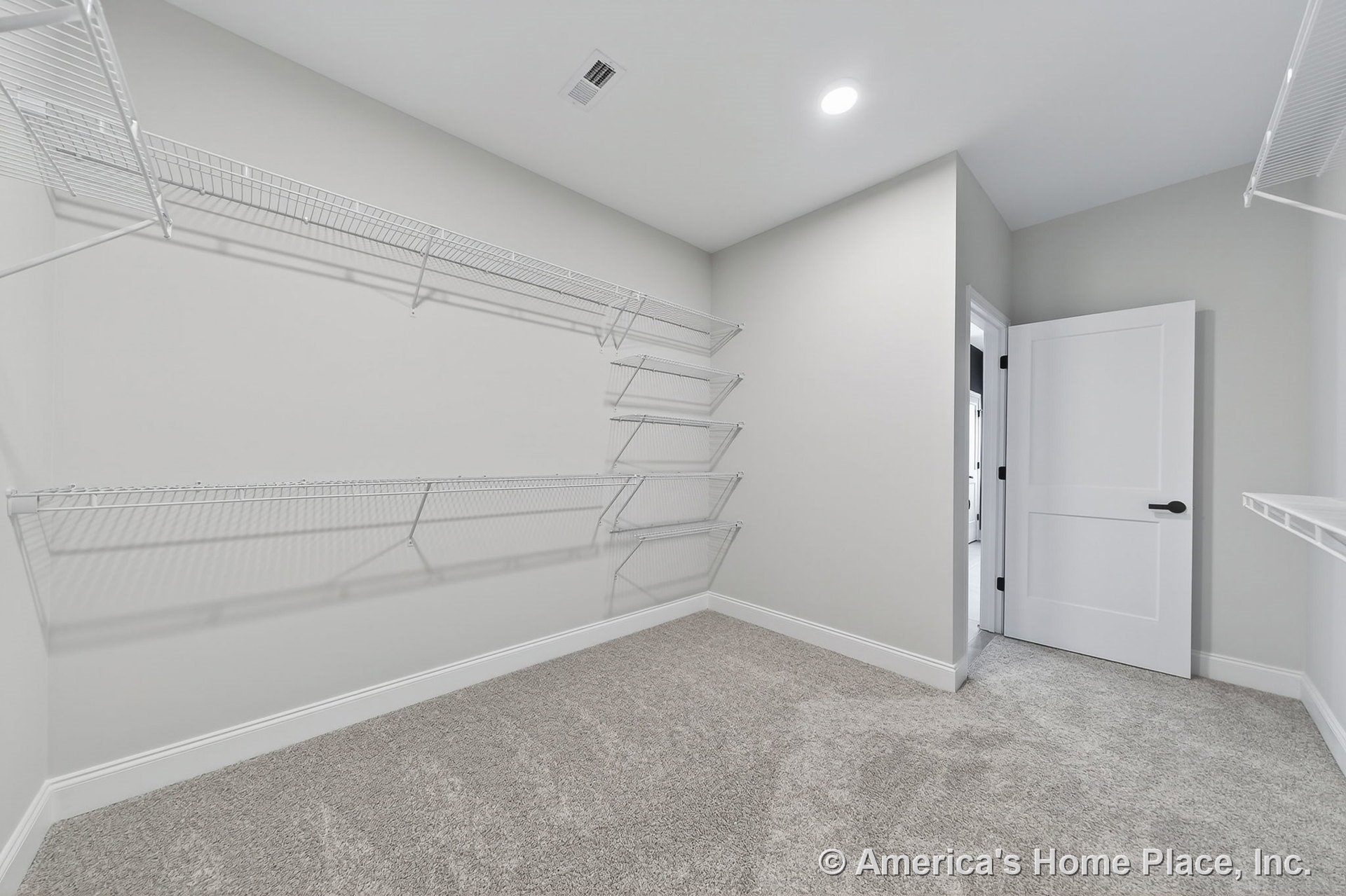 Walk-in closet with built-in white wire shelving, carpeted floor, recessed ceiling light, white trim and baseboards, panel door with black hardware, and neutral wall color.