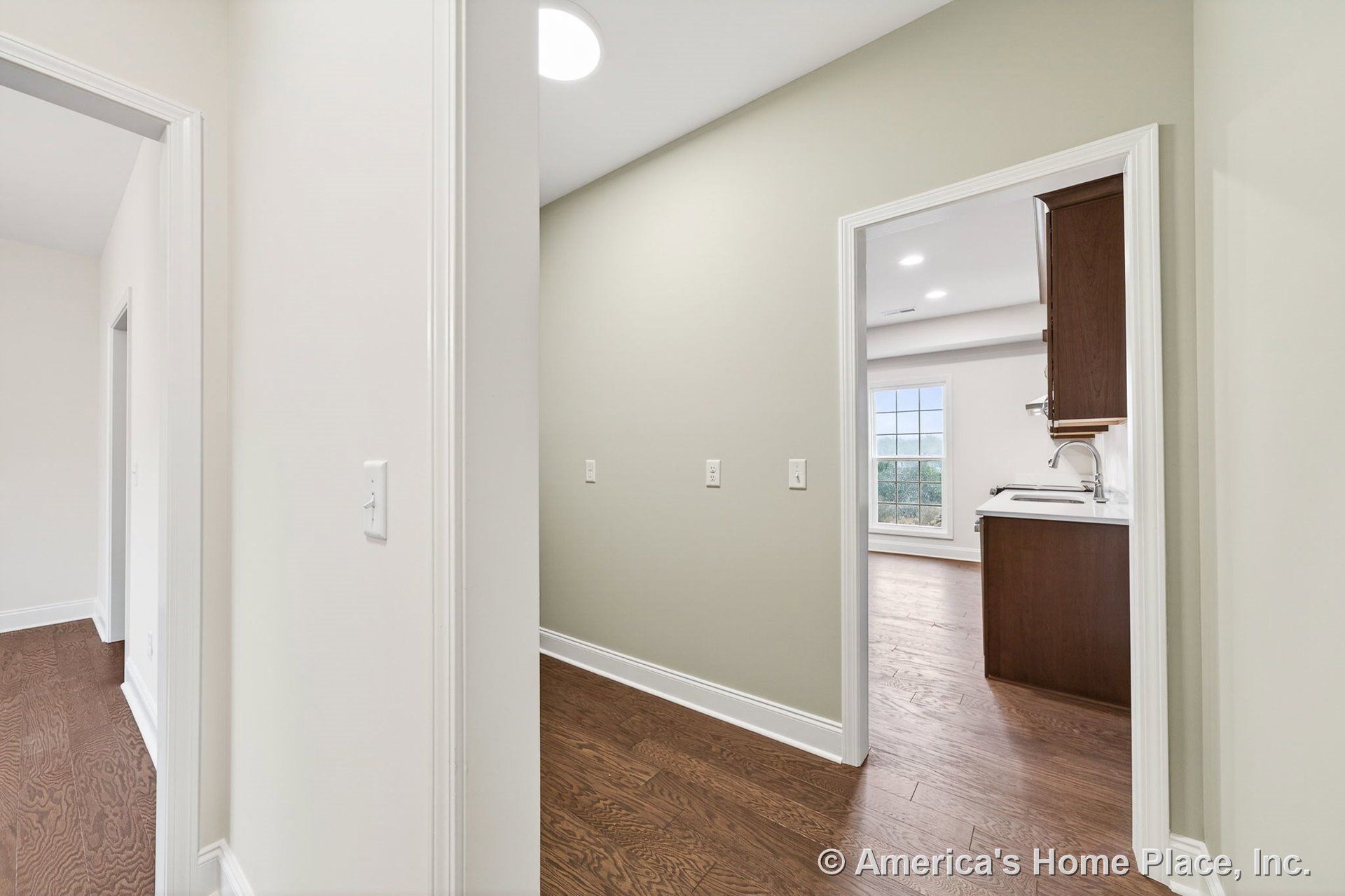Hallway with hardwood flooring, white trim and baseboards, recessed ceiling lighting, leading to kitchen area featuring built-in cabinetry with sink, large window, and neutral wall