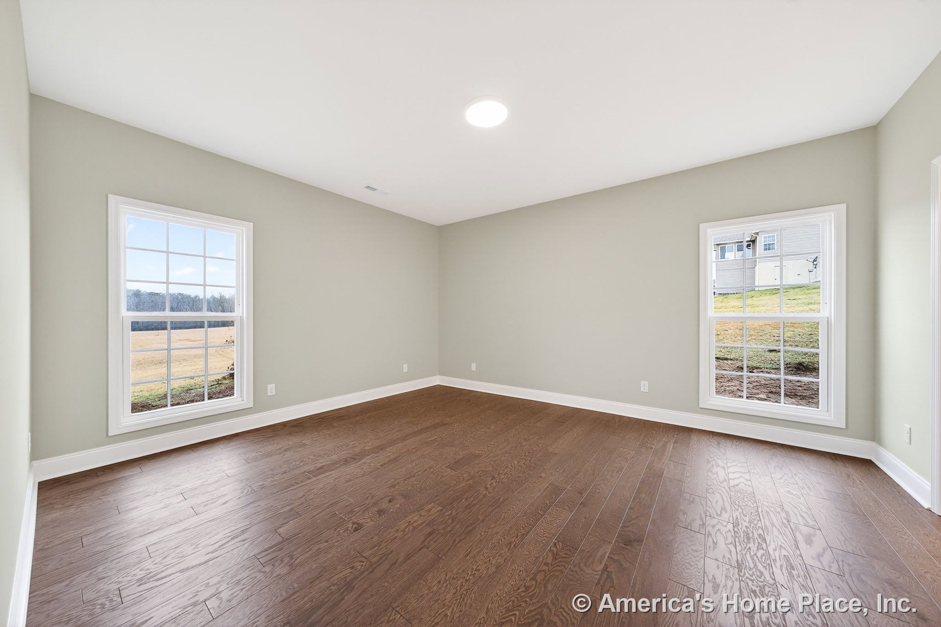 Wide plank hardwood flooring, two large double-hung windows with white trim, neutral wall color, recessed ceiling light, and baseboard molding in an empty modern bedroom.