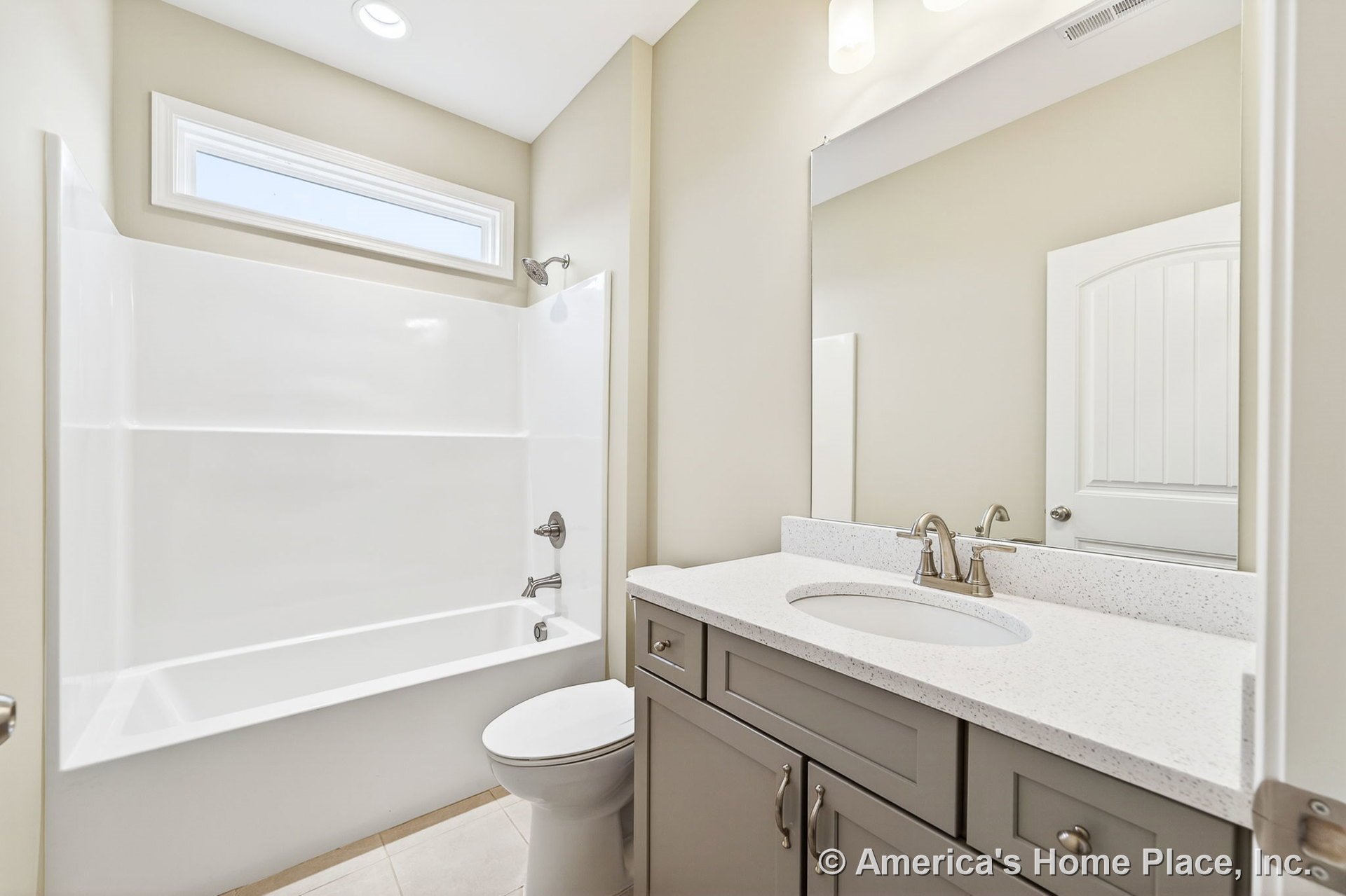 Built-in bathtub with shower fixtures beside a quartz countertop vanity featuring gray shaker cabinetry, large wall mirror, rectangular transom window, recessed ceiling light, tile